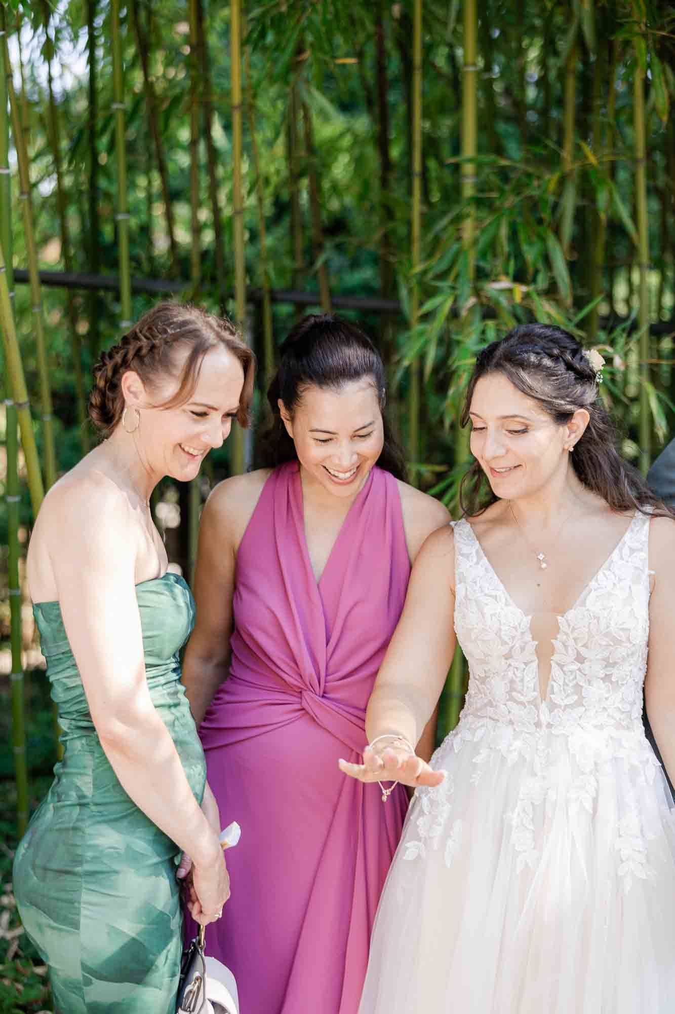 Bride in white lace gown laughing with bridesmaids in green and purple dresses outdoors near bamboo.