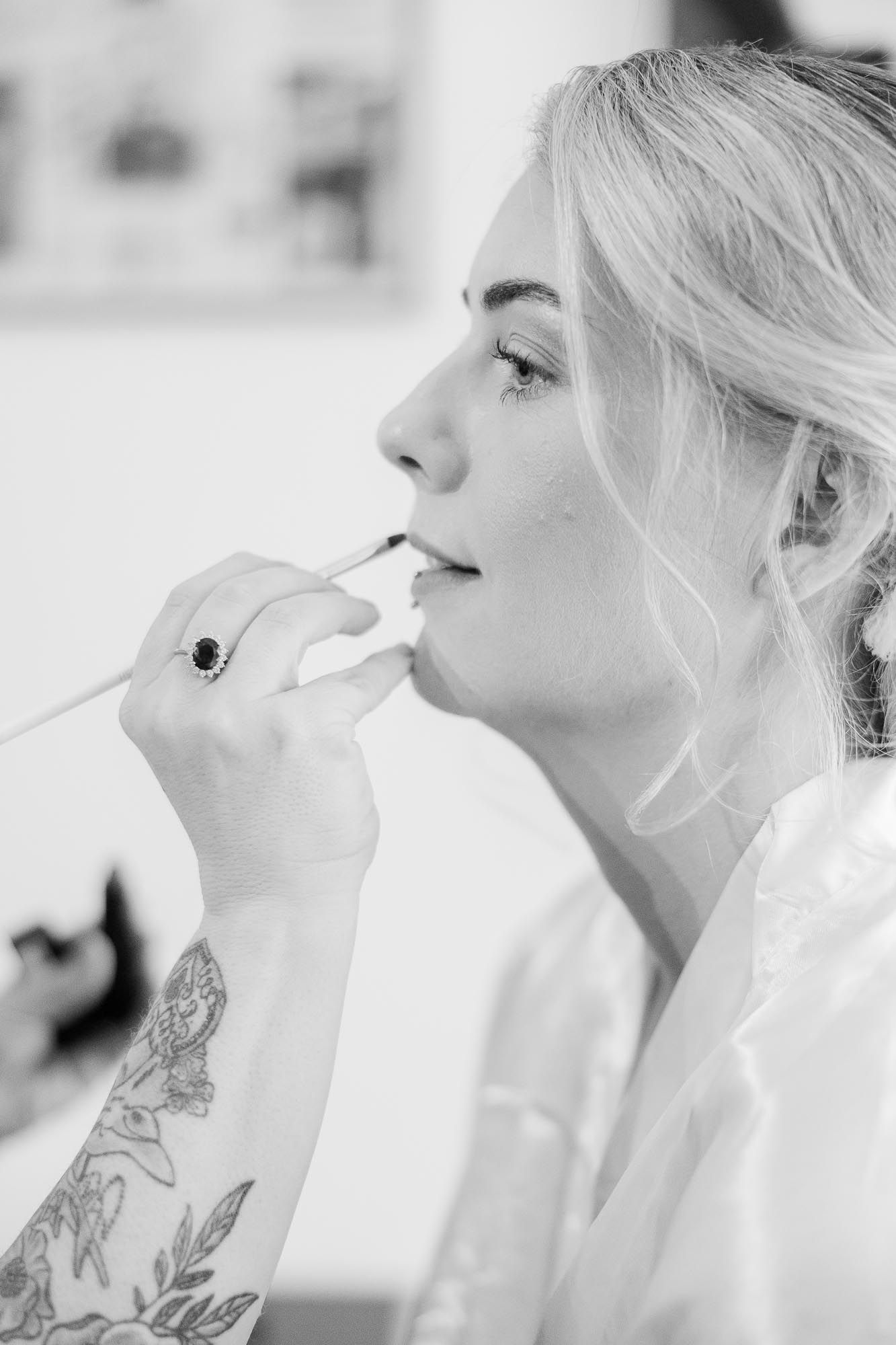 Black and white portrait of blonde bride applying lipstick while getting ready for wedding.
