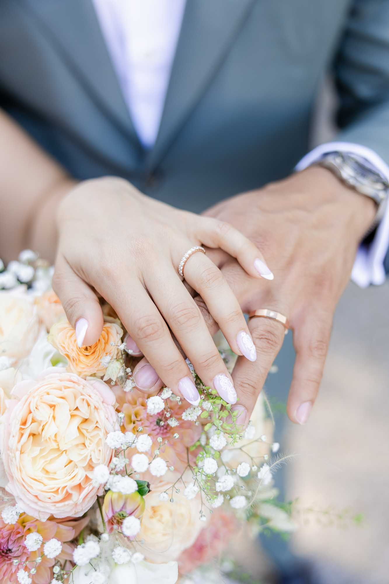 Newlyweds displaying gold wedding rings on hands resting over a bridal bouquet of peach roses and white flowers.