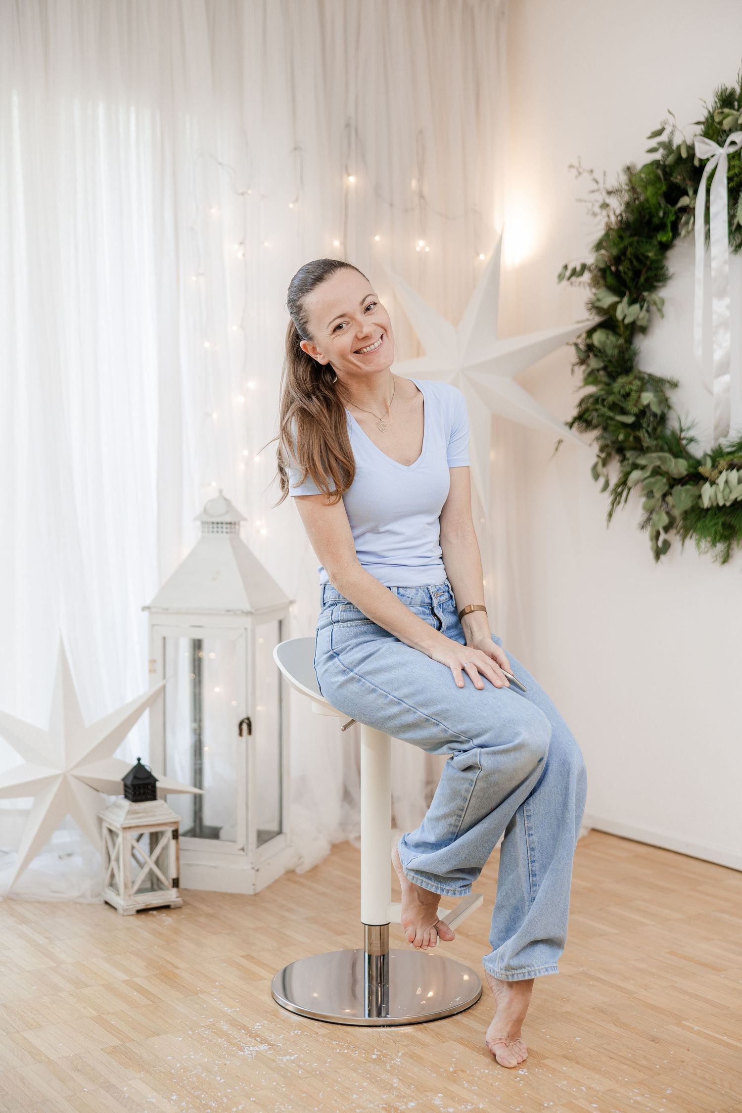 Woman in gray tank top and light denim overalls poses playfully on wooden floor with white decor and string lights.