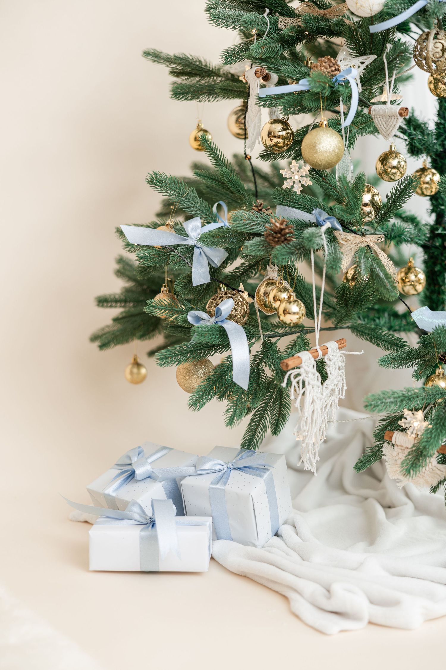 Decorated Christmas tree with gold ornaments, blue ribbons and white wrapped presents underneath on white blanket.
