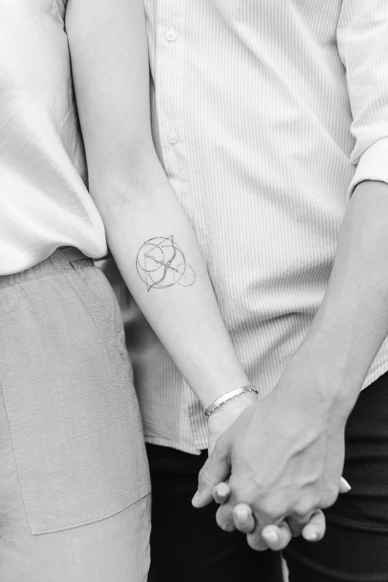 Black and white photo showing clasped hands and a small circular geometric tattoo on someone's forearm. Couple shoot in Switzerland