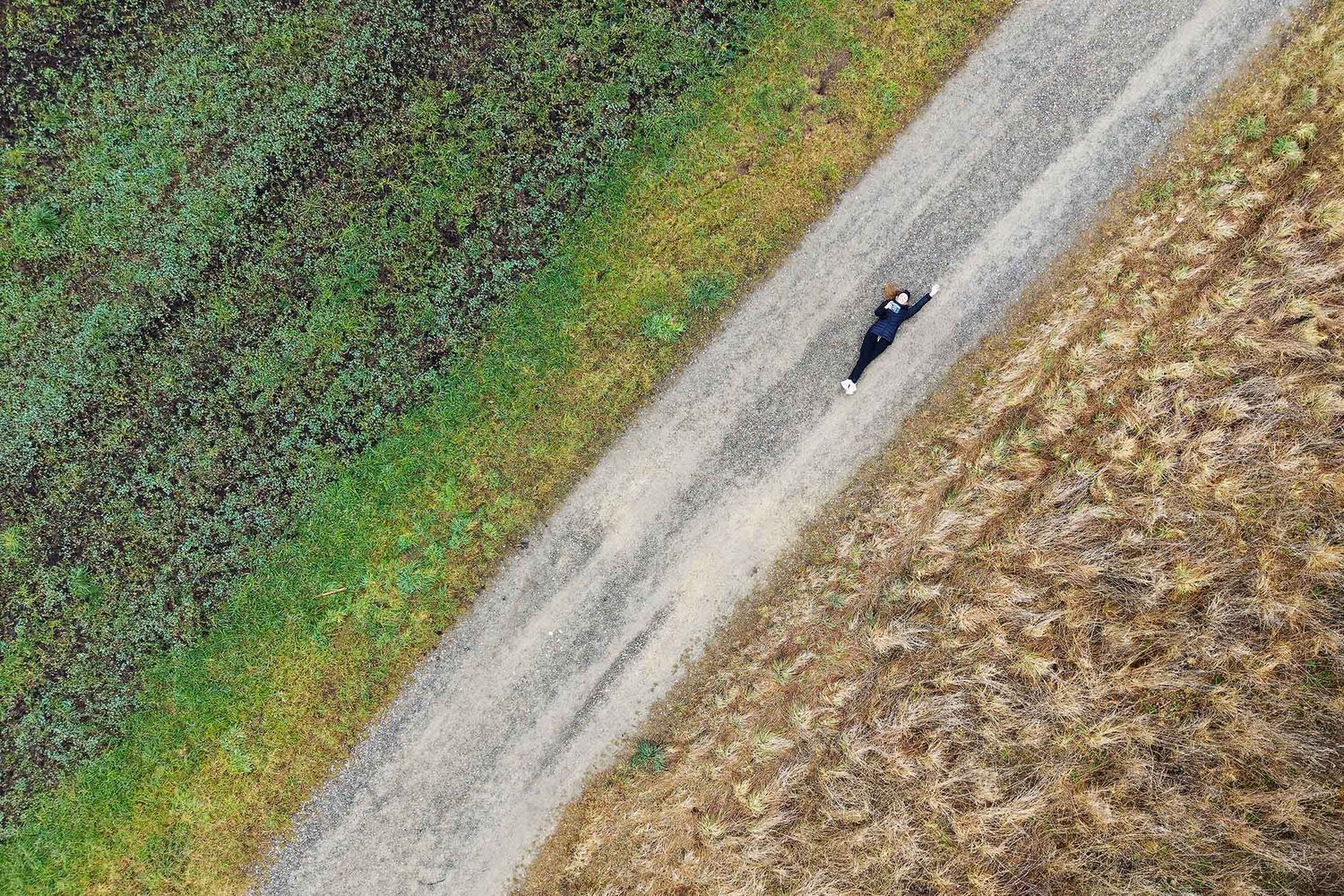 Aerial view of a lone black car driving on a rural dirt road between green vegetation and brown harvested field.