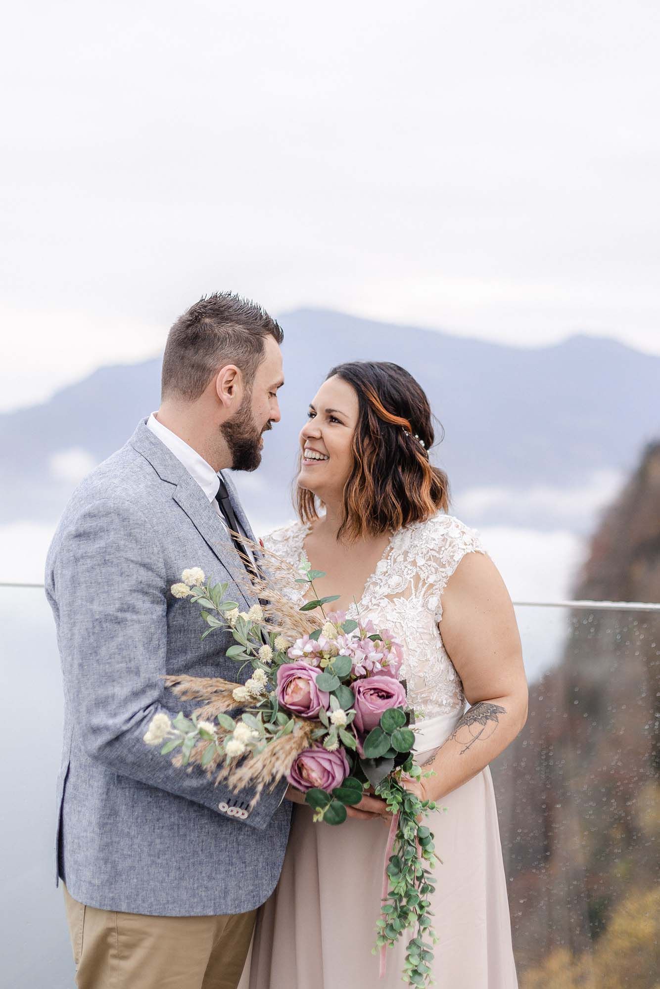 A couple shares a joyful moment on their wedding day against a misty mountain backdrop with a cascading floral bouquet.
