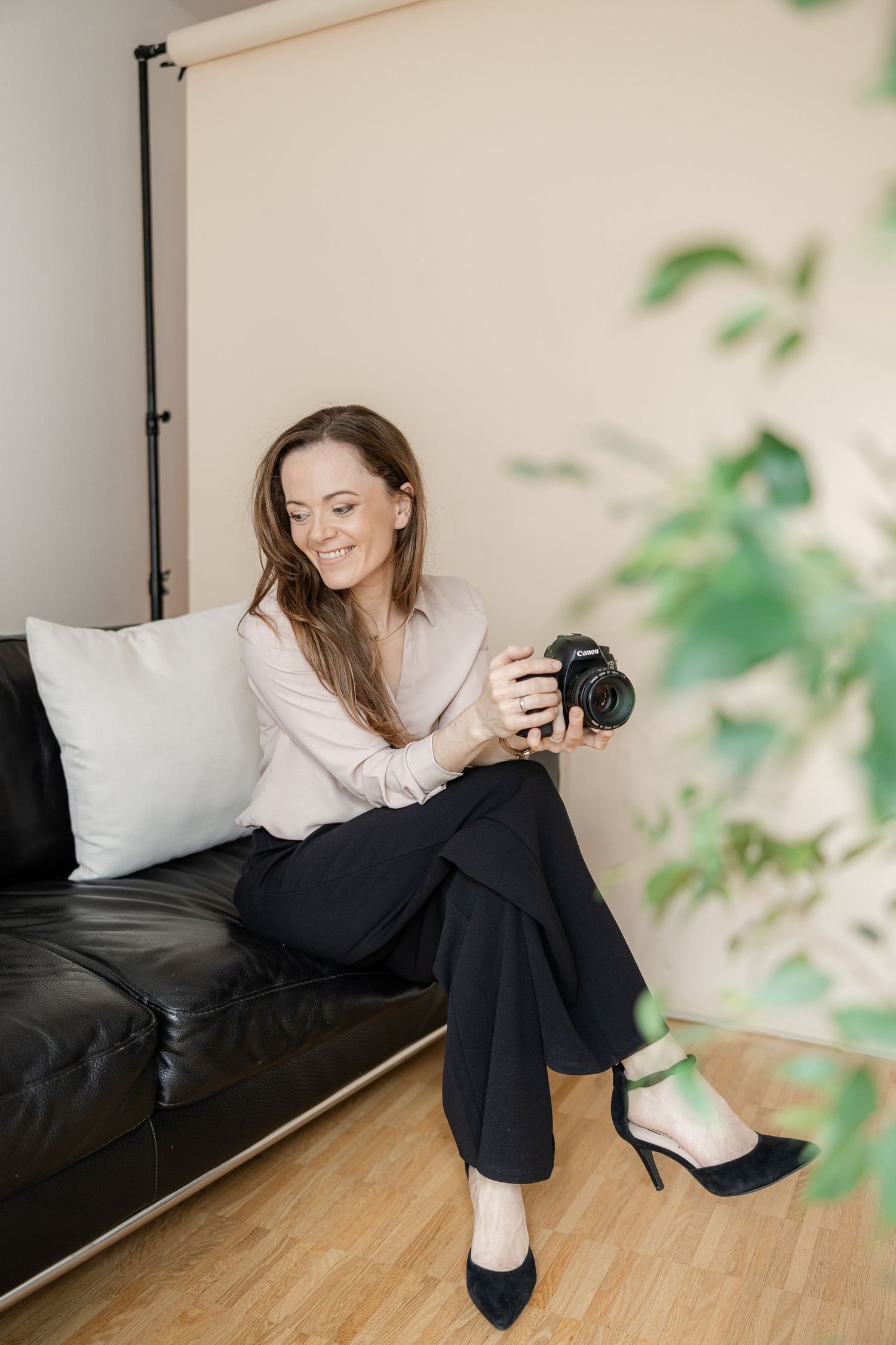 A photographer wearing a beige sweater and black pants sits on a leather couch while holding a professional camera.