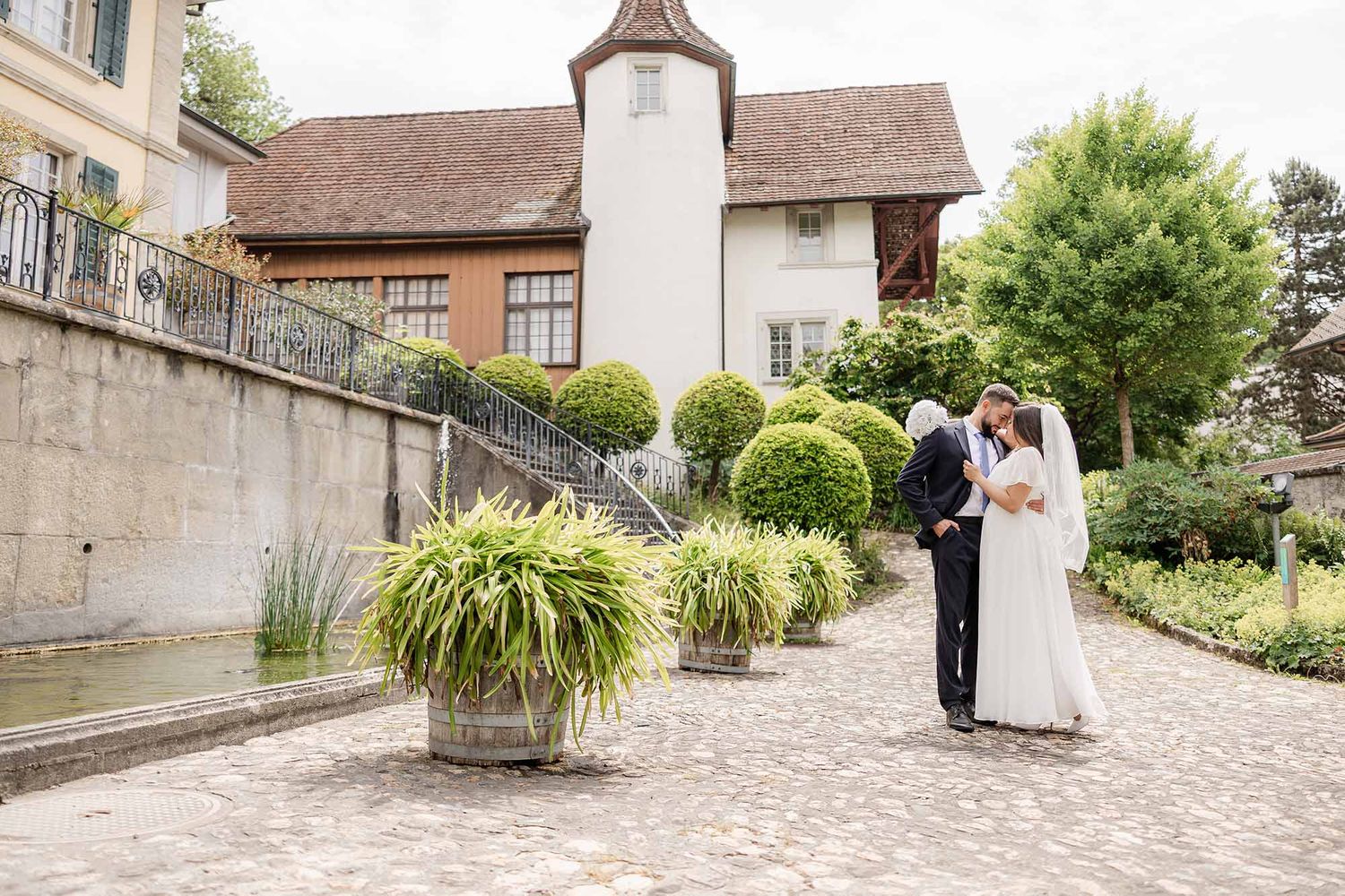 Bride and groom share a romantic moment in an elegant European garden courtyard with manicured topiary and stone architecture.