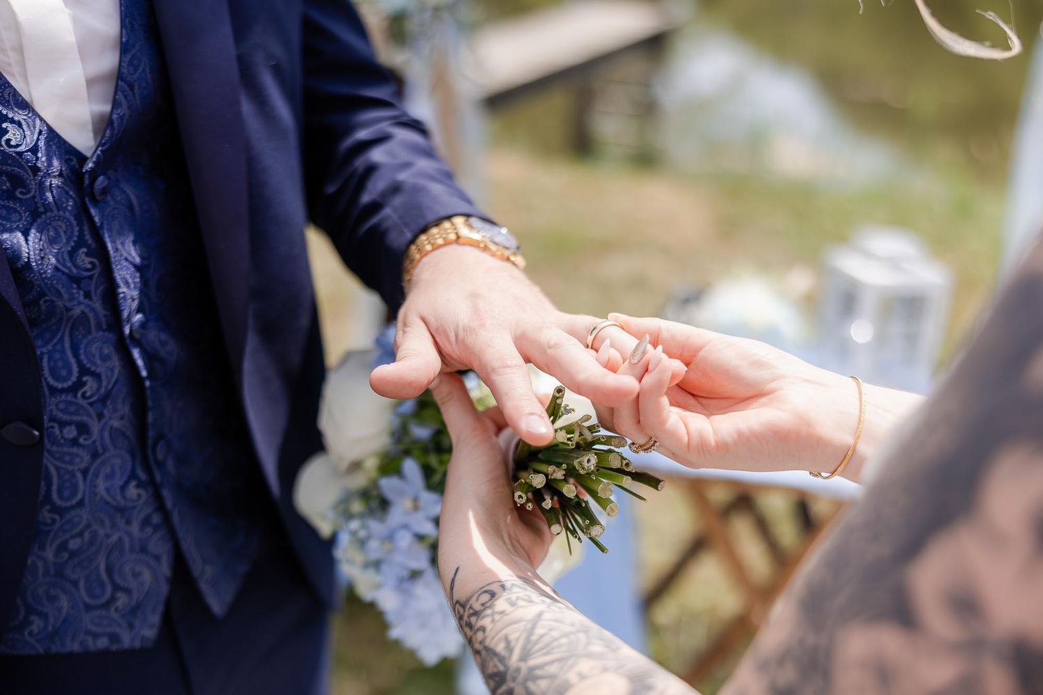 Close-up of a wedding ring exchange, groom in blue suit with tattoo placing ring on bride's finger.
