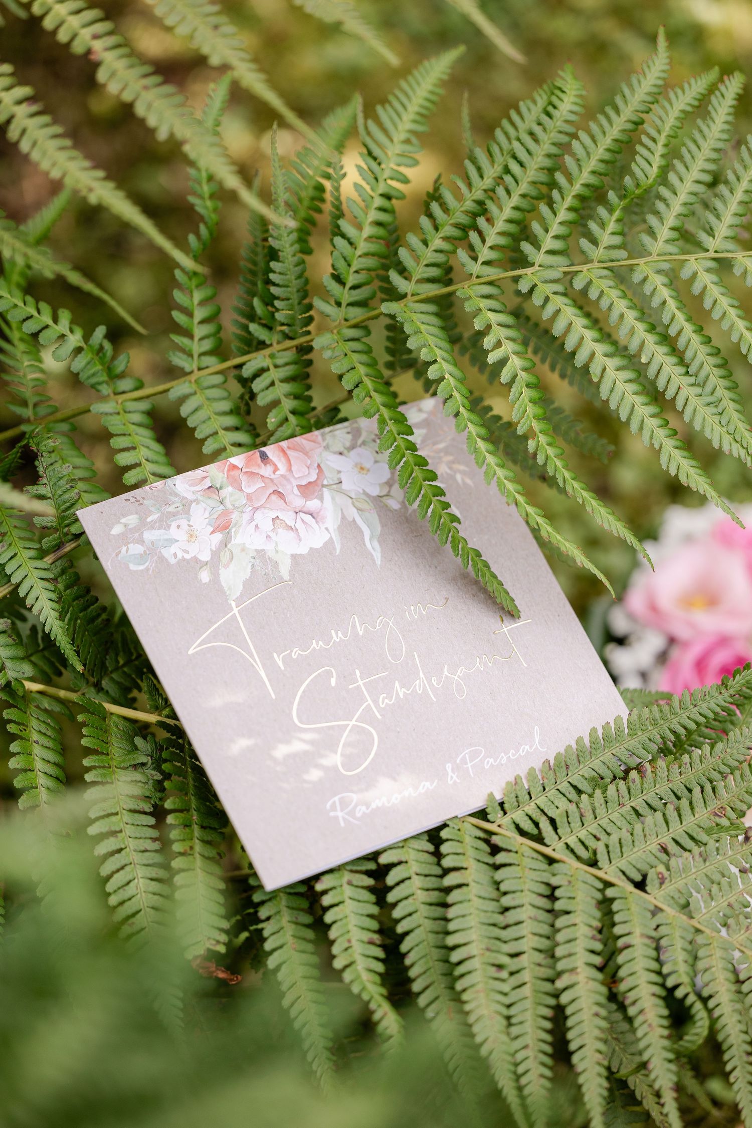 Wedding invitation cards displayed on green fern leaves with delicate calligraphy and floral designs.