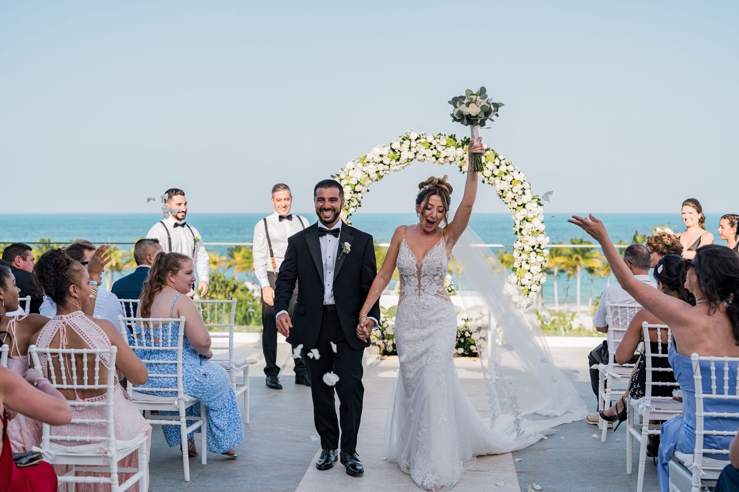 Joyful bride raises bouquet while walking down aisle with groom at outdoor beach wedding ceremony.