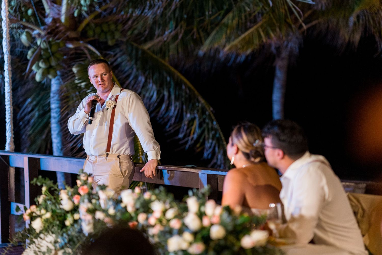 Man giving speech at elegant outdoor beach wedding reception with floral centerpieces and palm trees.