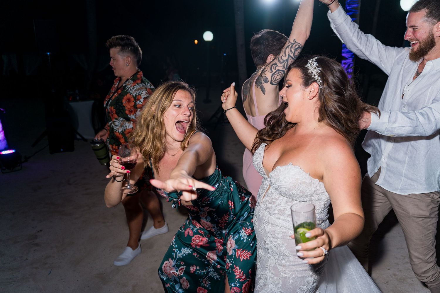 Bride in strapless gown celebrates with laughing guests on a sandy beach wedding dance floor.