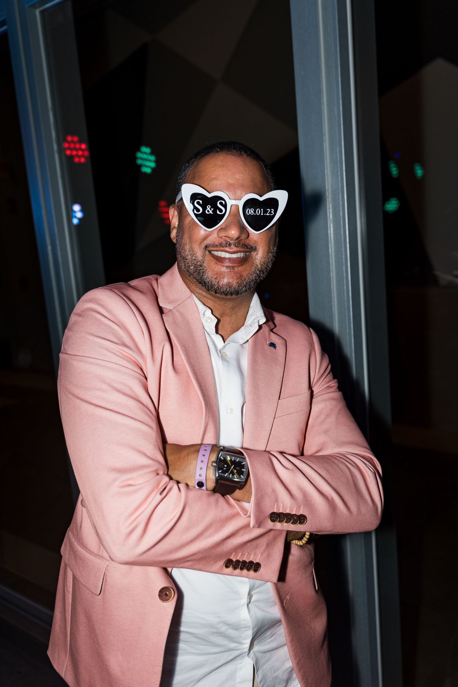 Man in pink blazer wearing heart-shaped S&S wedding sunglasses poses with arms crossed at night.