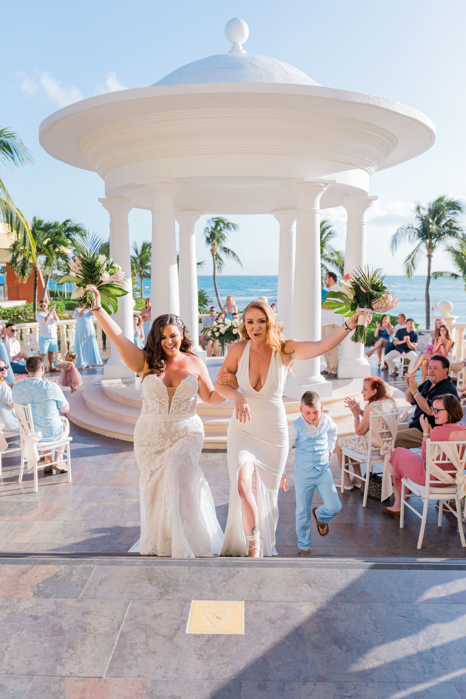 Two brides celebrating joyfully with raised arms after their tropical oceanfront wedding ceremony.