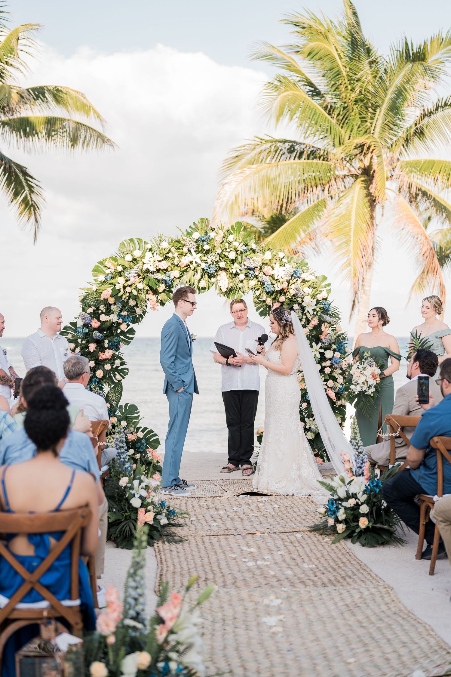 Bride and groom exchange vows under floral arch at tropical beach wedding ceremony at sunset.