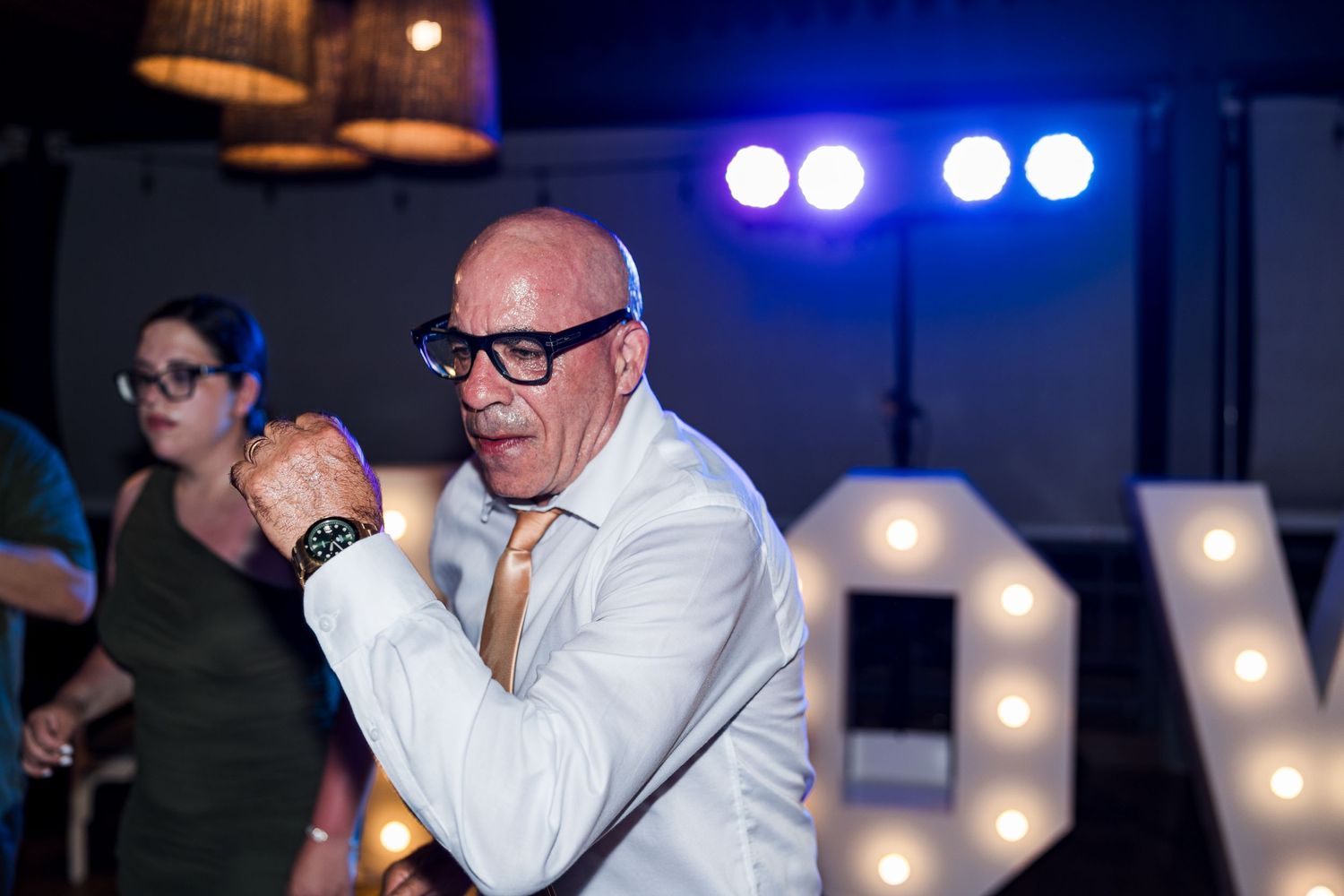 Energetic older man dancing enthusiastically in front of illuminated LOVE letters at wedding reception.