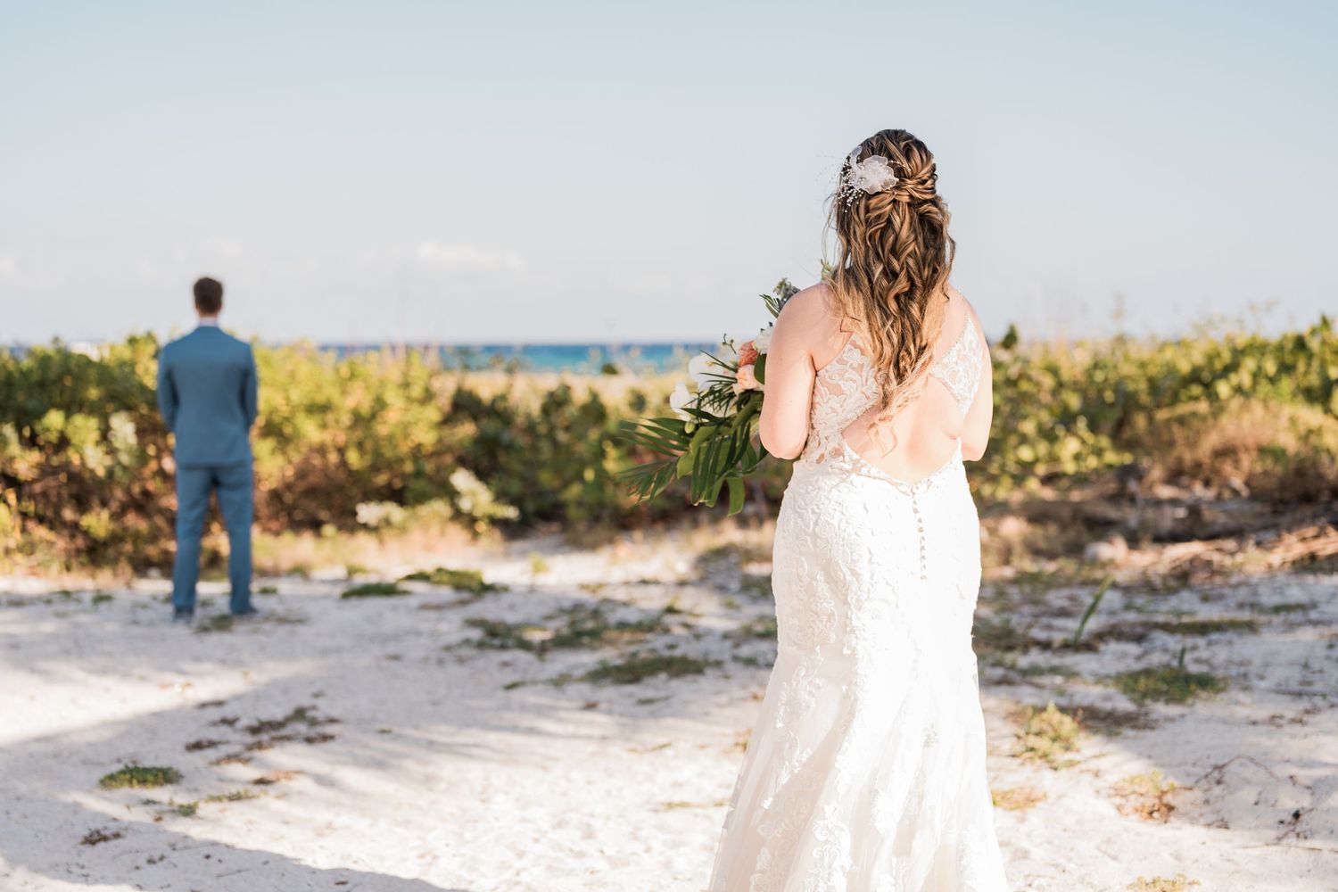 Bride in lace gown holding bouquet approaches groom for first look on sandy beach.