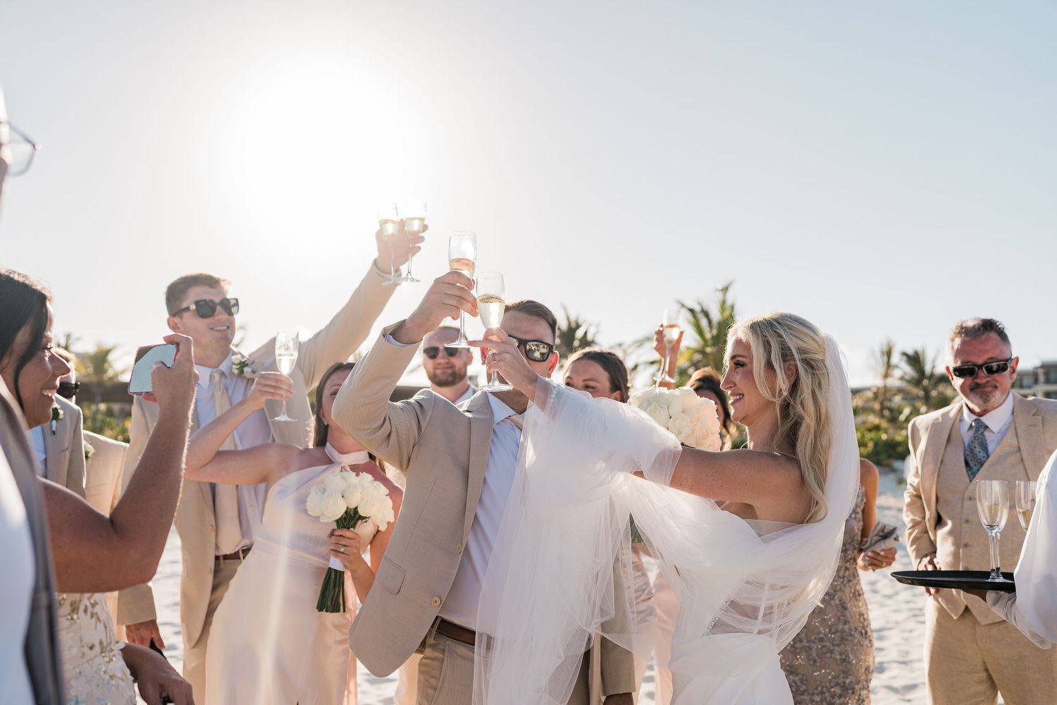 Newlyweds and wedding party raise champagne glasses in joyful beach celebration under bright sunny sky.