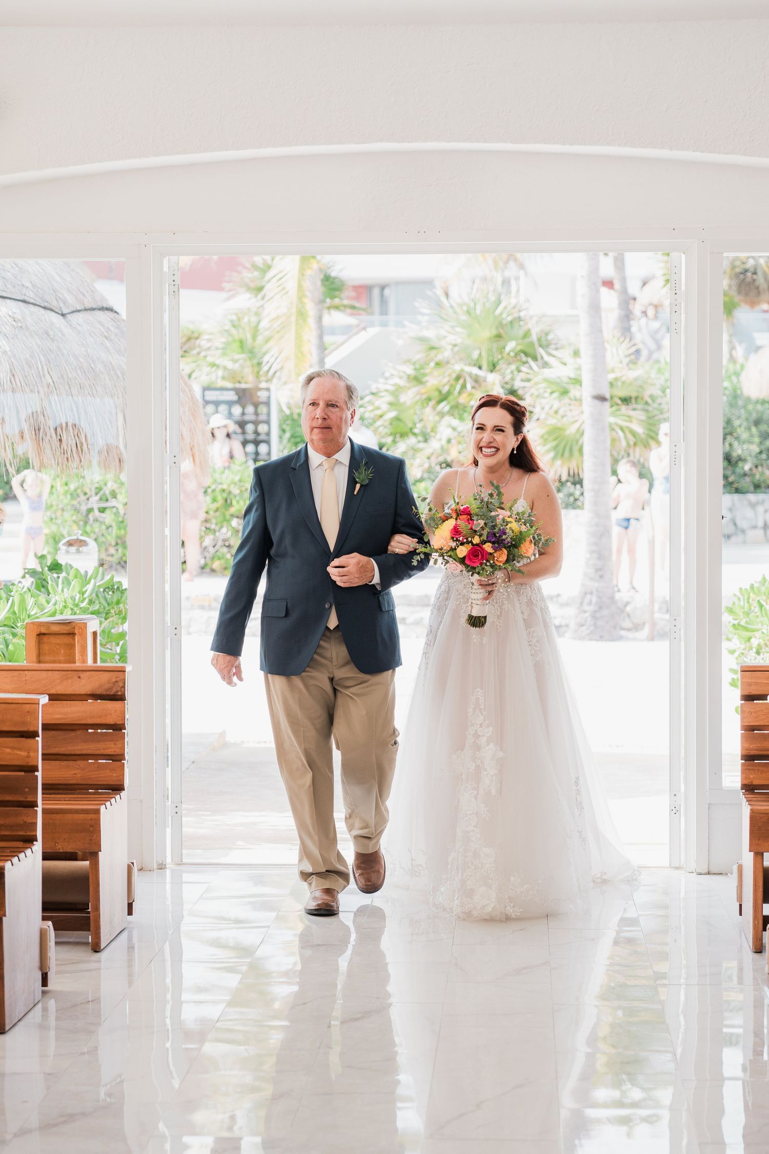 Smiling bride walks down chapel aisle with father holding colorful bouquet at destination wedding.