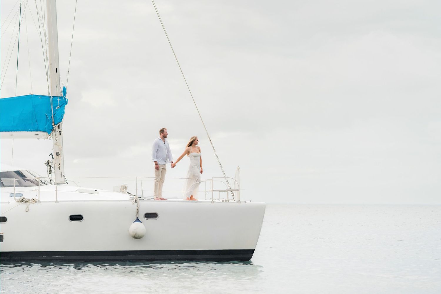 Couple holding hands on a white sailboat catamaran on calm ocean waters under overcast skies.