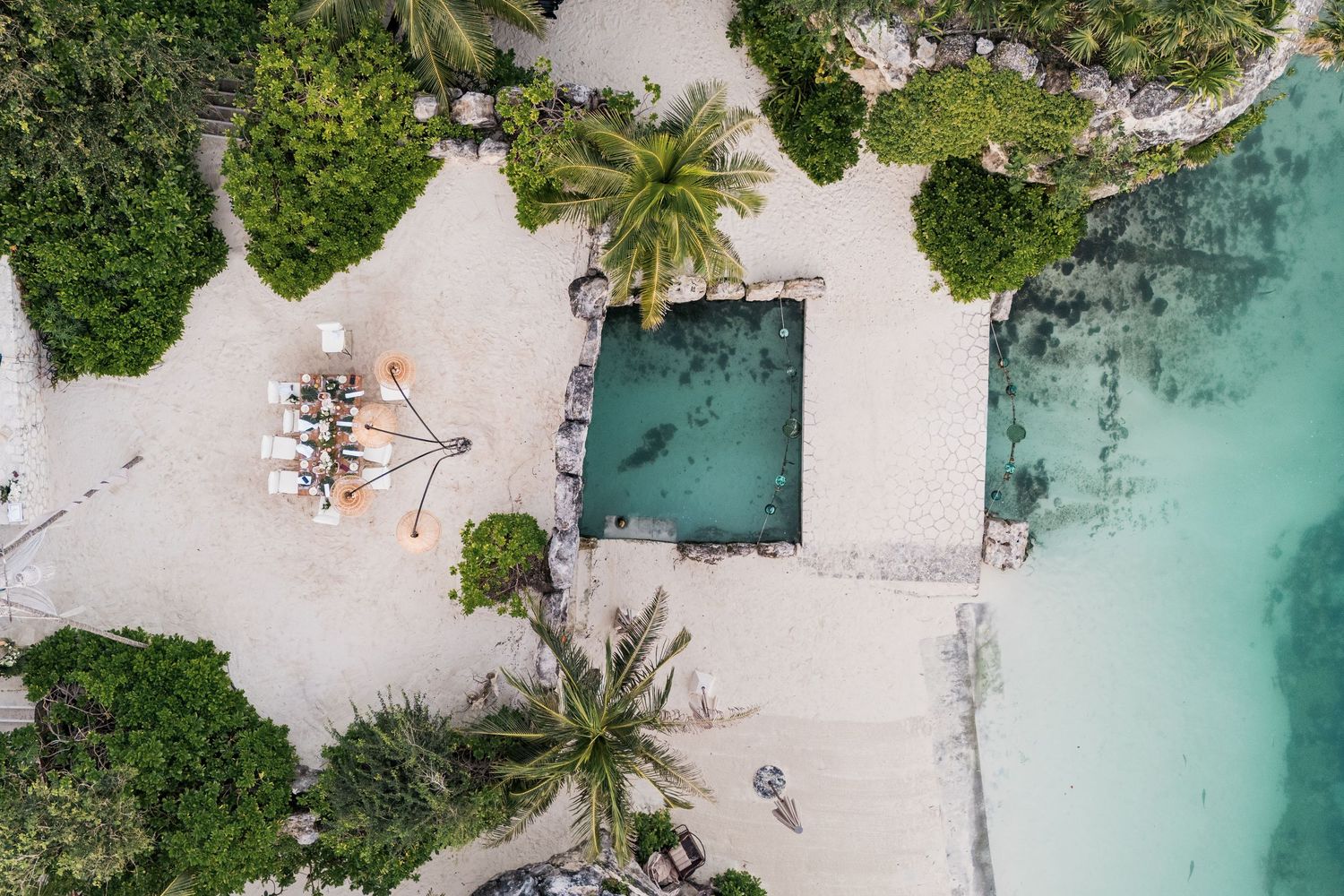 Xcaret Arte Wedding - Aerial view of a tropical beach resort with a rectangular pool, white sand, lounge chairs, and turquoise ocean waters.