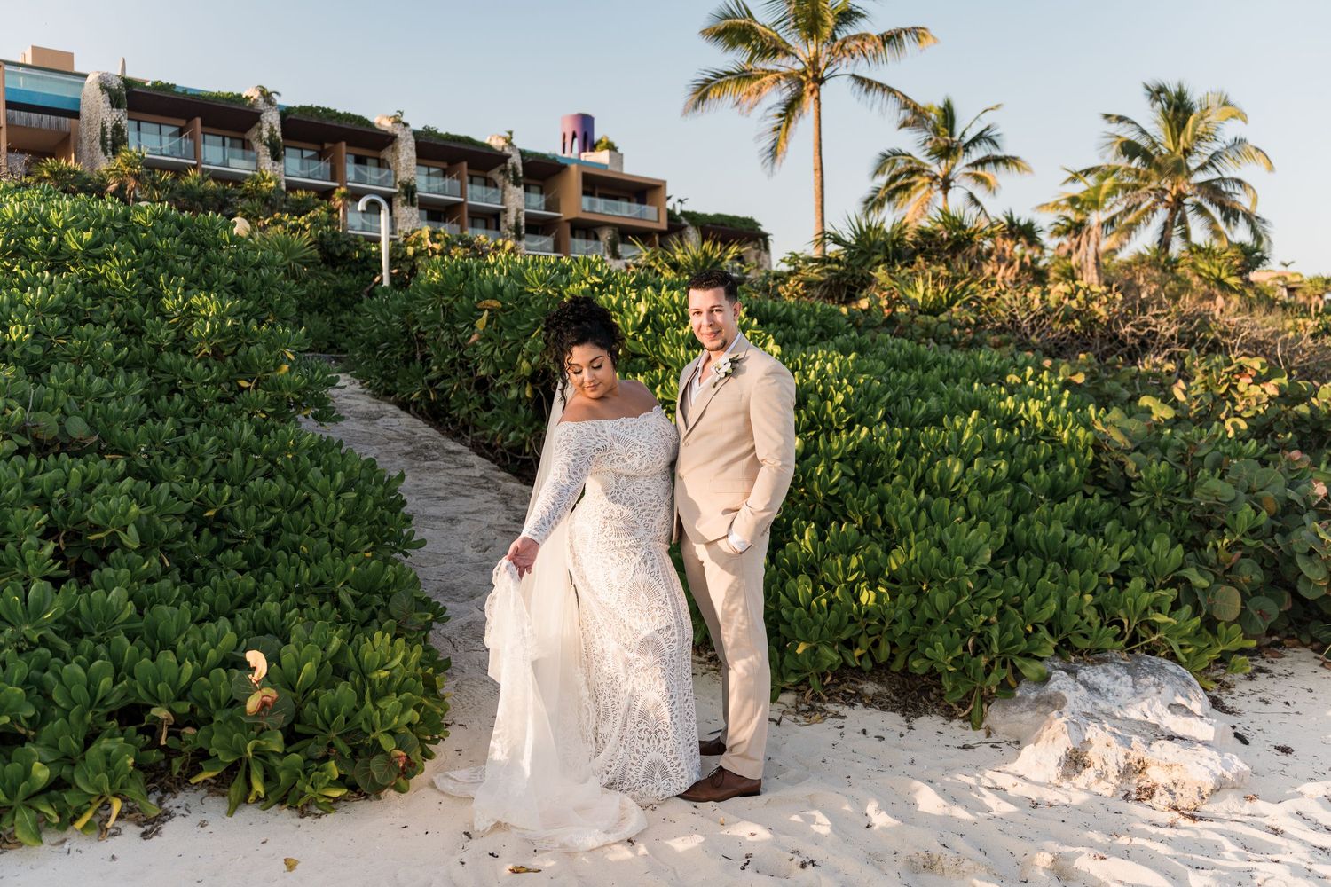 Xcaret Arte Wedding - Bride and groom posing on a sandy beach path surrounded by tropical greenery at golden hour.
