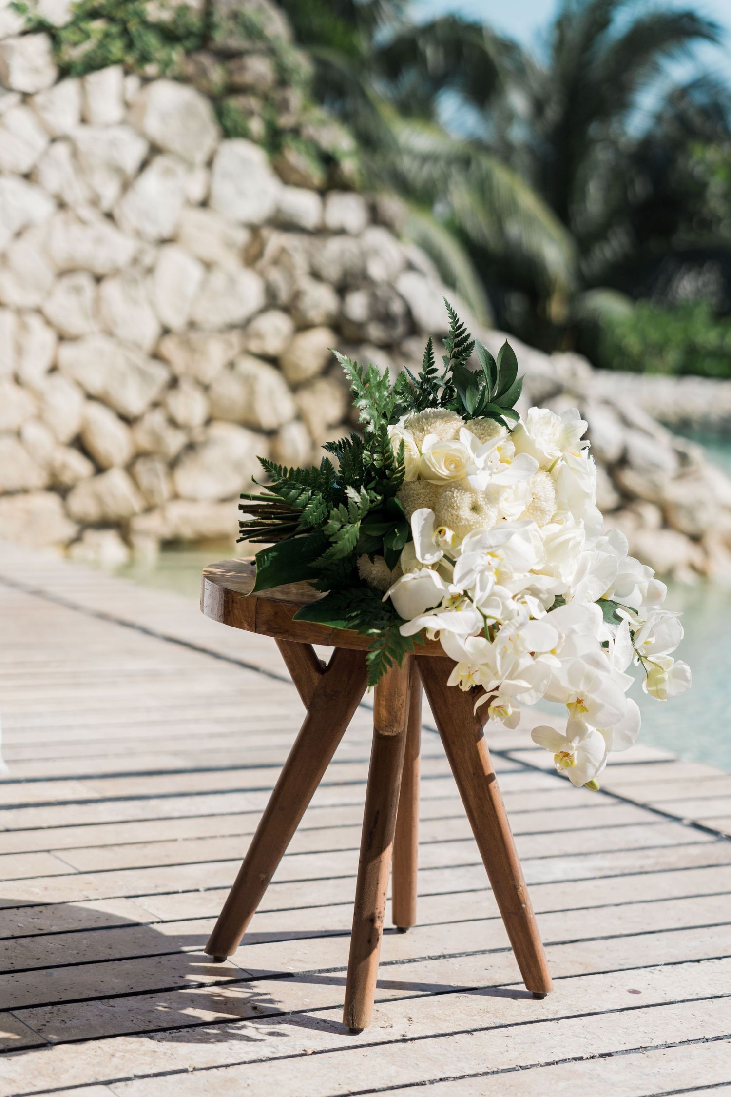 Xcaret Arte Wedding - White orchid and fern bouquet on wooden stool by tropical pool with stone wall backdrop.