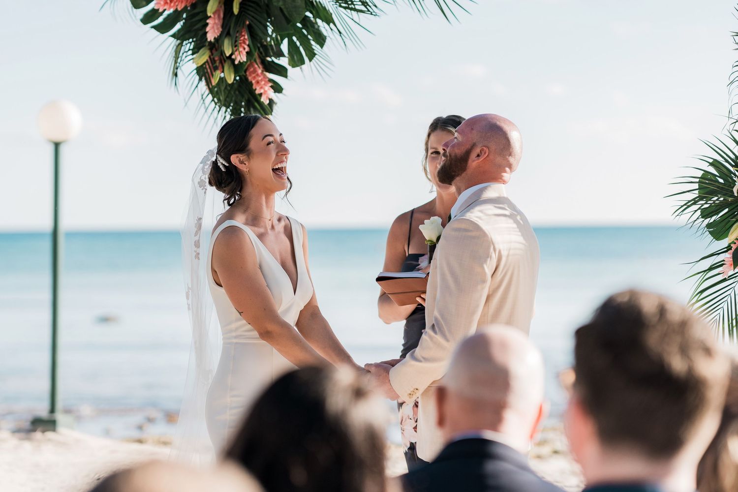 Bride and groom exchange vows at a tropical beach wedding ceremony with guests seated and palm trees in the background.