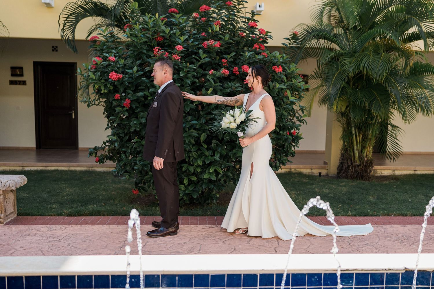 Bride and groom share a first look moment by a pool, surrounded by lush greenery and red flowers at an outdoor venue.