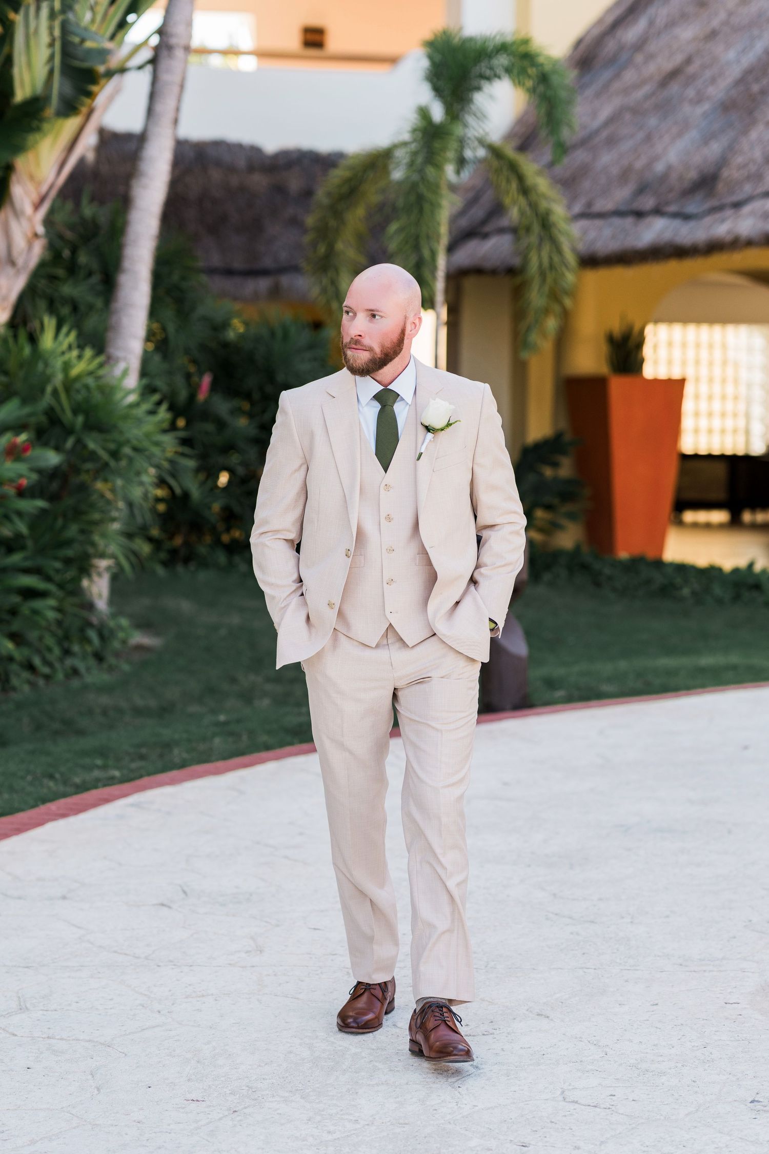 Groom in a beige three-piece suit with green tie and boutonniere stands confidently outdoors at a tropical wedding venue.