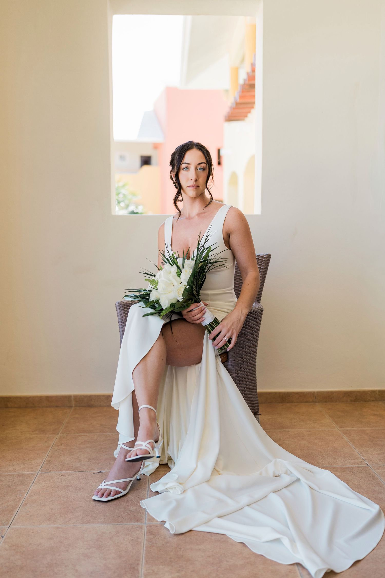Elegant bride in ivory gown holding white floral bouquet, seated in wicker chair by window in warm sunlit room.