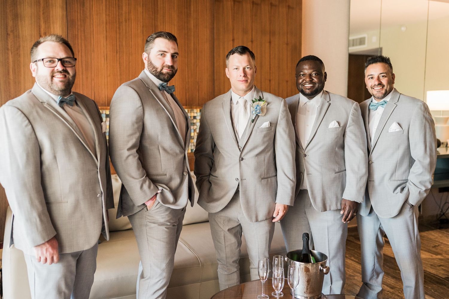 Five groomsmen in matching gray suits with bow ties smiling together indoors at grand palladium costa mujeres wedding