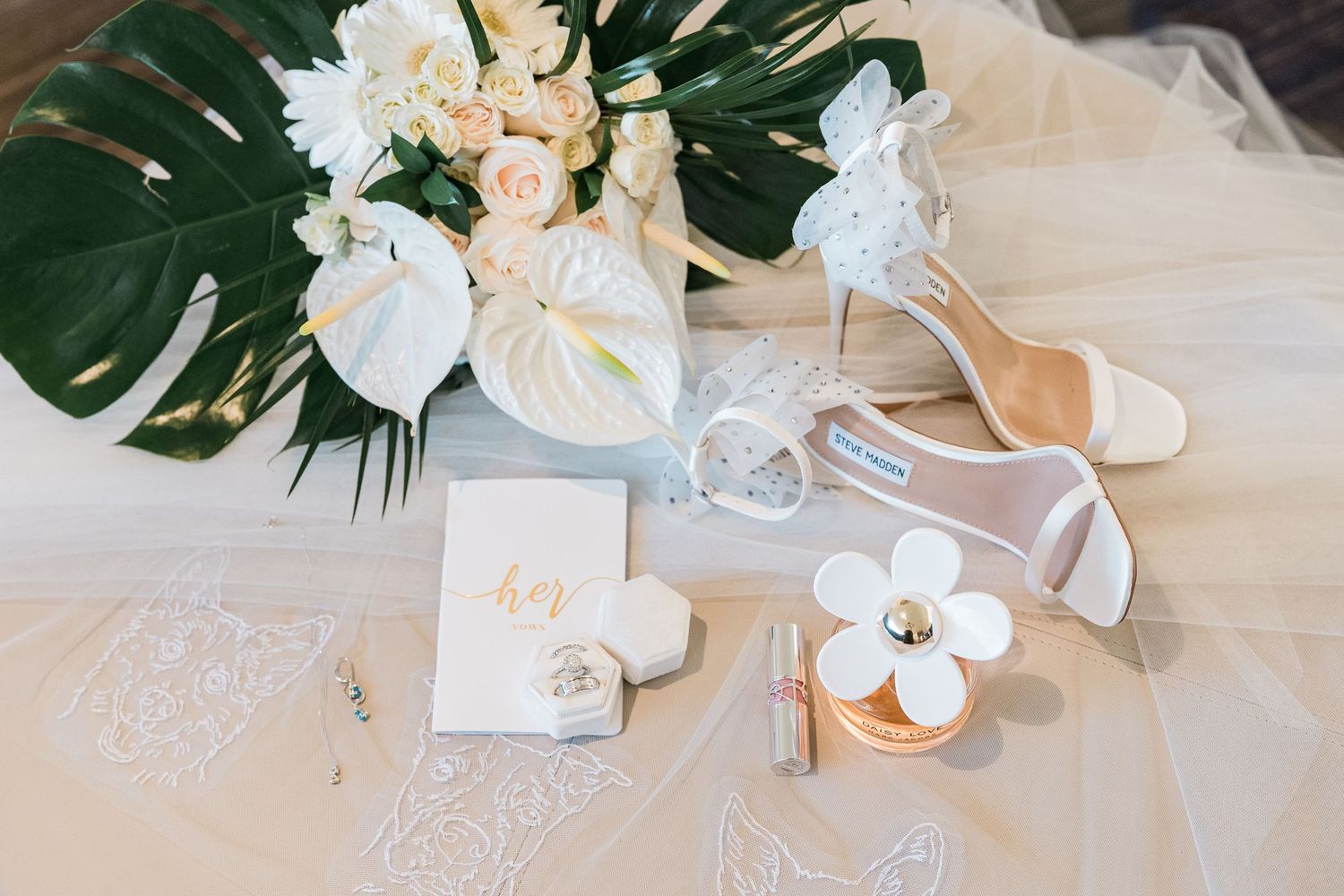 Elegant bridal flat lay with white heels, floral bouquet, veil, gold stationery, and perfume on a soft white surface.