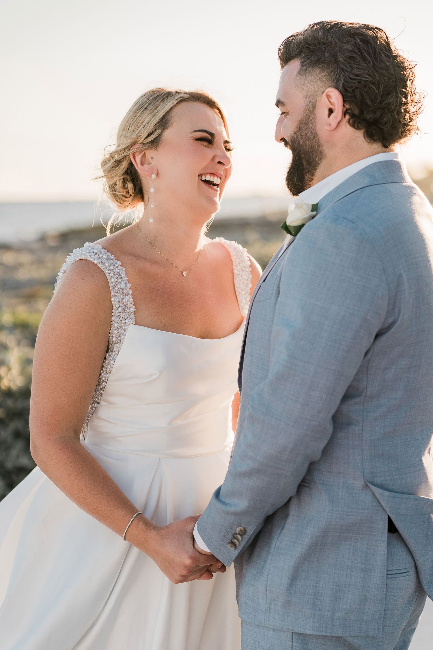 Bride in white gown laughs with bearded groom in light blue suit during golden hour wedding portrait outdoors.