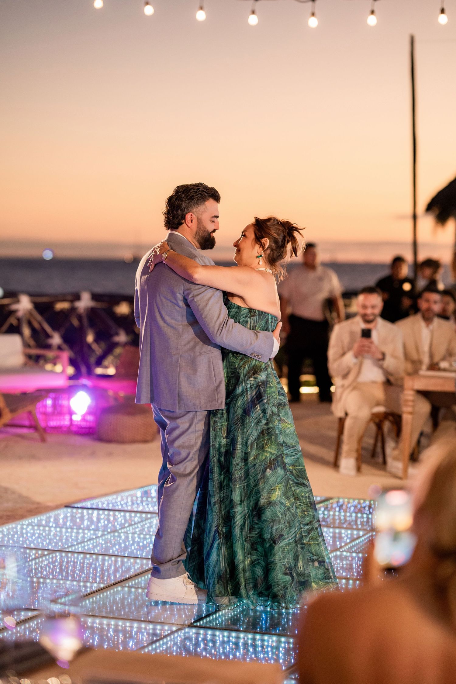 Couple sharing first dance on illuminated floor at beach wedding reception during golden hour sunset.