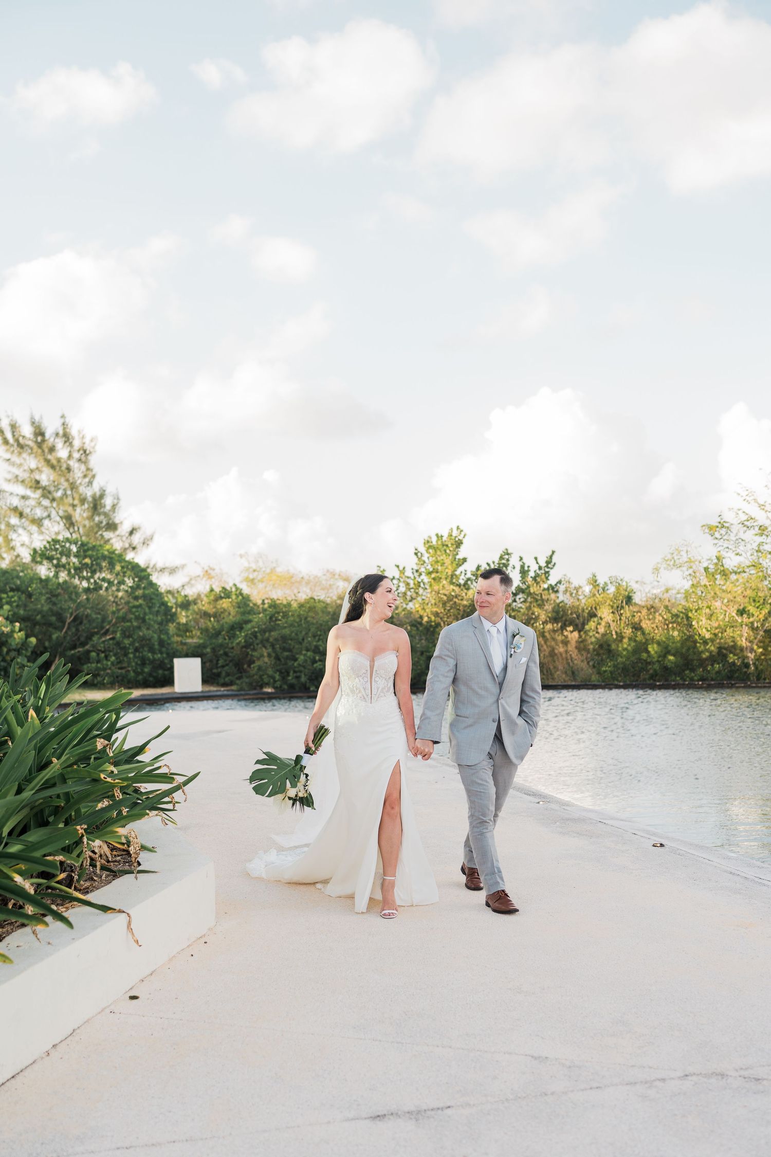 Bride and groom walk hand-in-hand in tropical setting, she in fitted white gown, he in light gray suit.