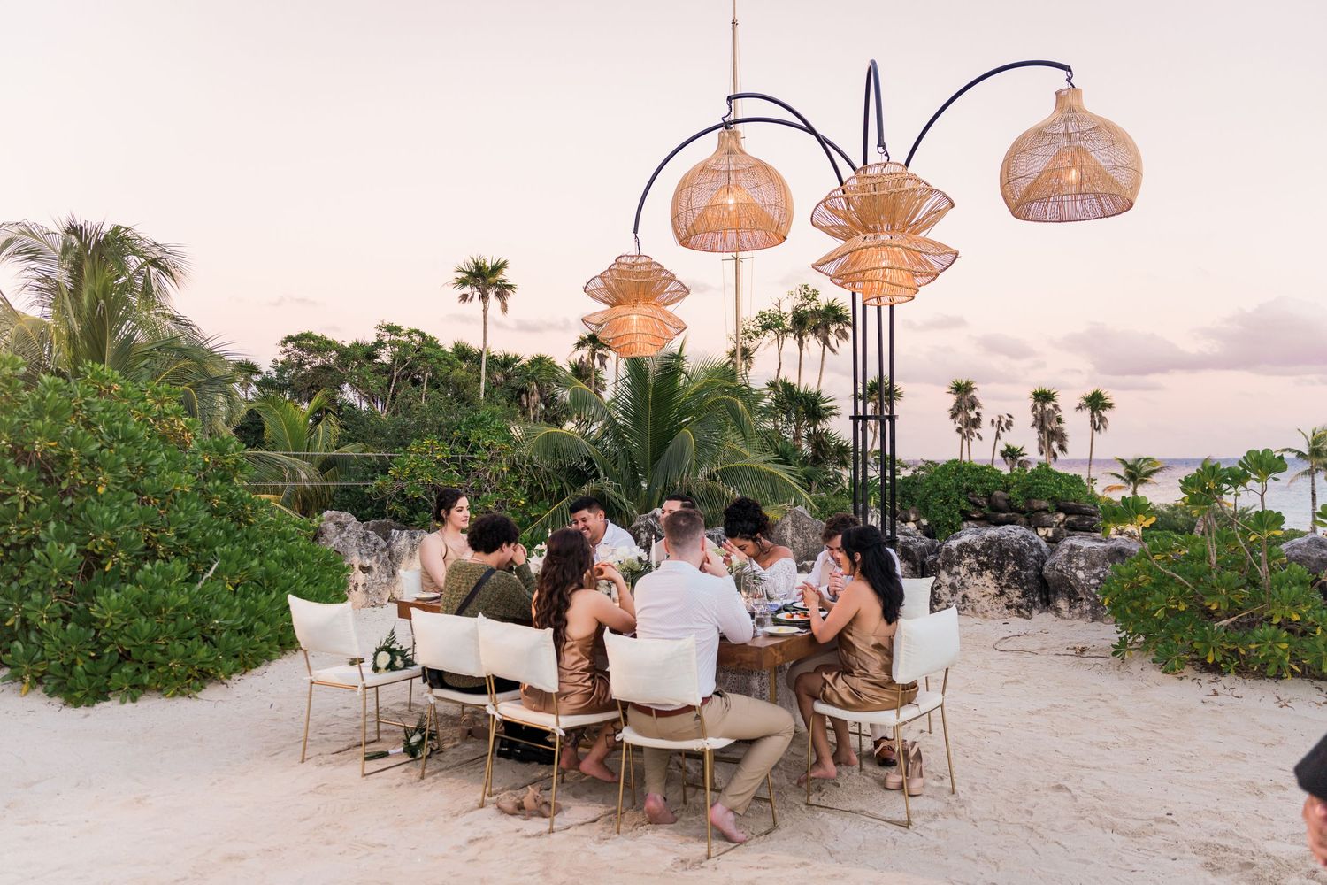 Group dining on sandy beach at sunset with wicker pendant lights, surrounded by tropical palms in a boho outdoor setting.