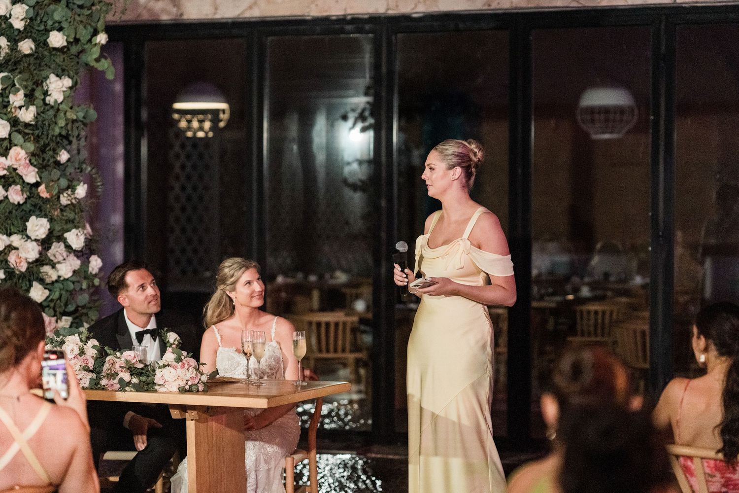 Woman in yellow dress giving a speech at an elegant wedding reception with floral decorations.