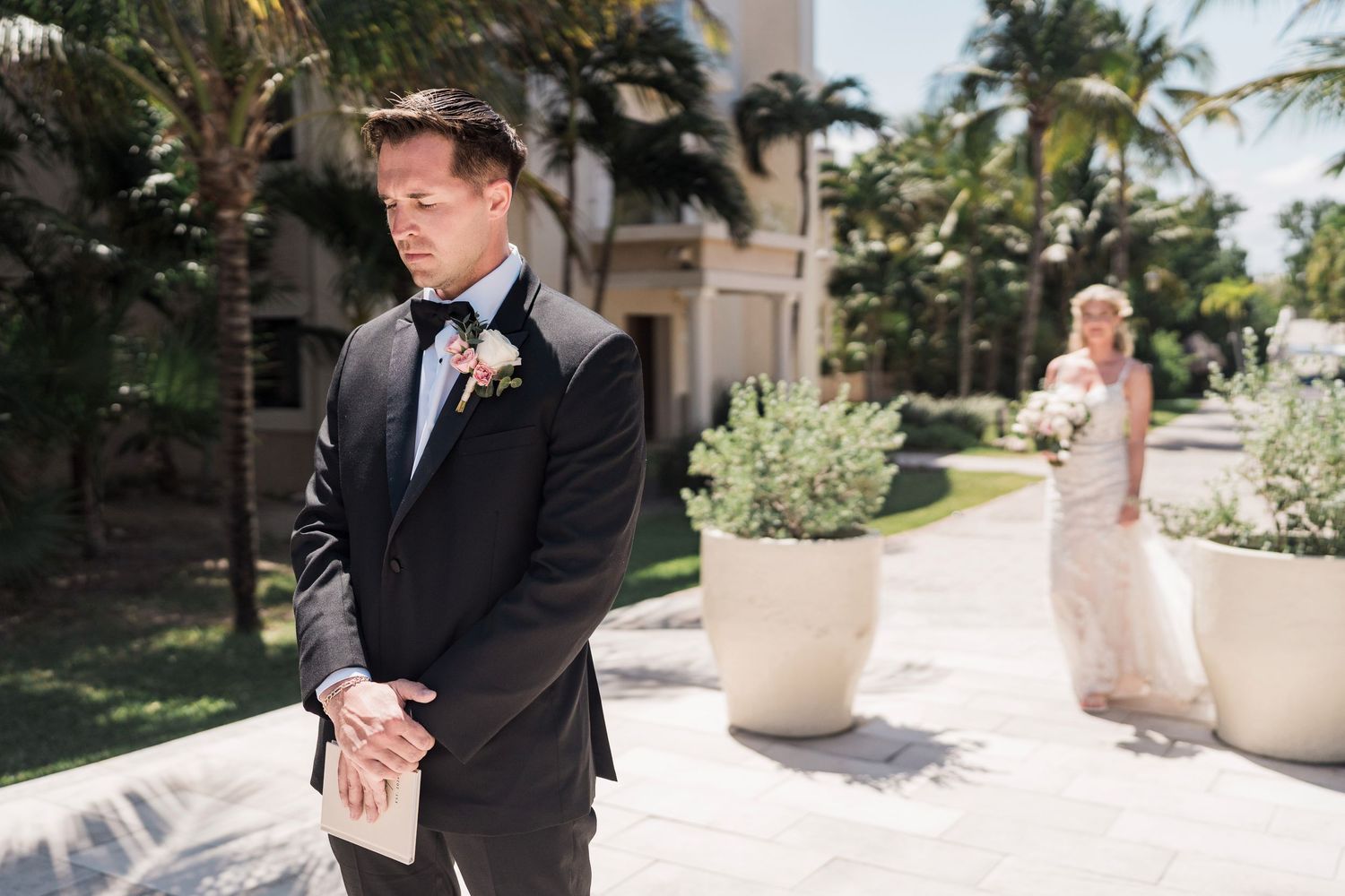 Groom in black tuxedo awaits bride in white gown for first look at tropical outdoor wedding venue.