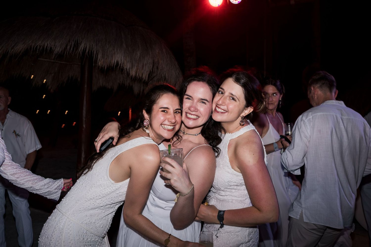 Three women in white dresses smiling and hugging at an outdoor evening party.