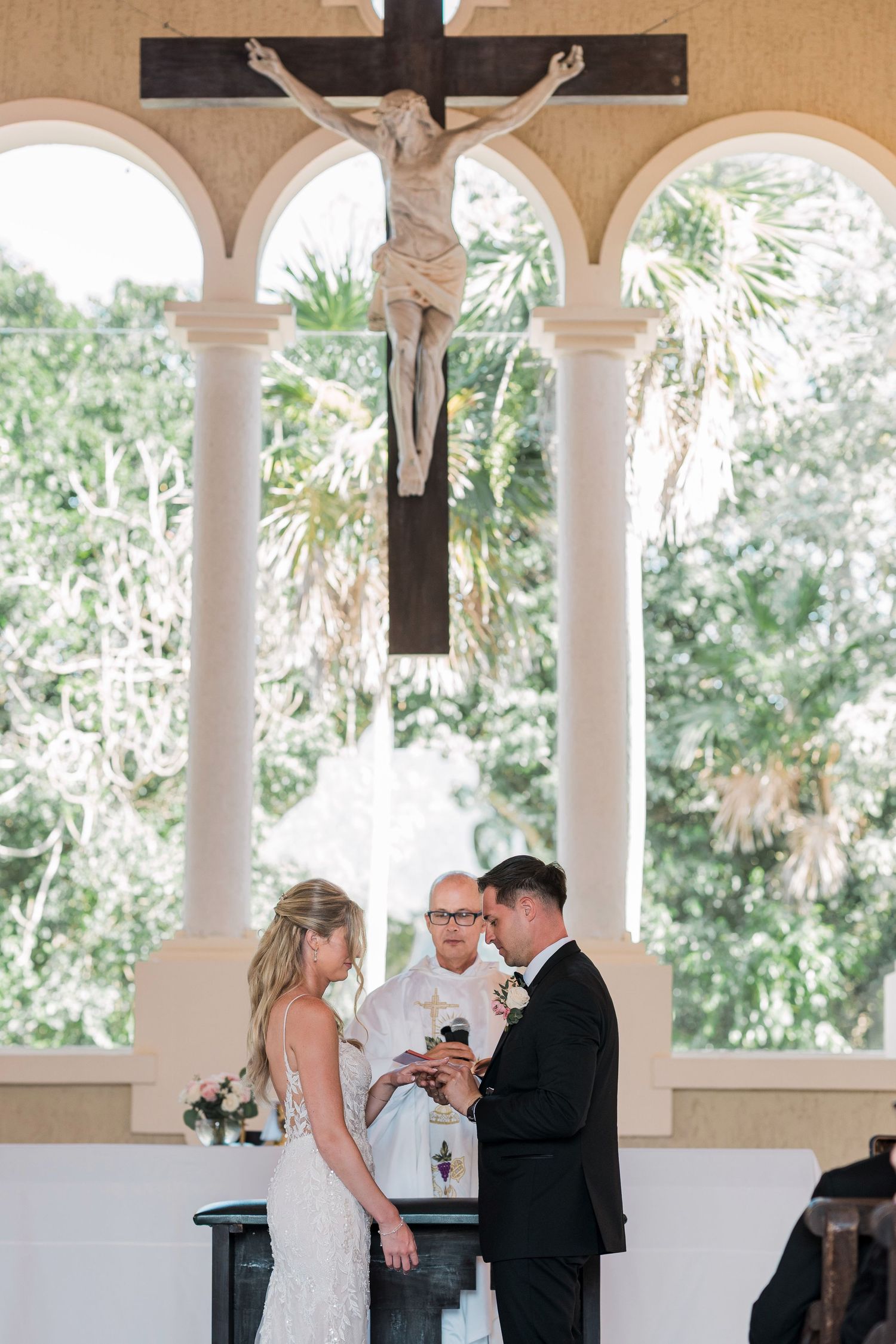 Bride and groom exchange vows beneath a crucifix at an outdoor Catholic wedding ceremony with lush greenery.
