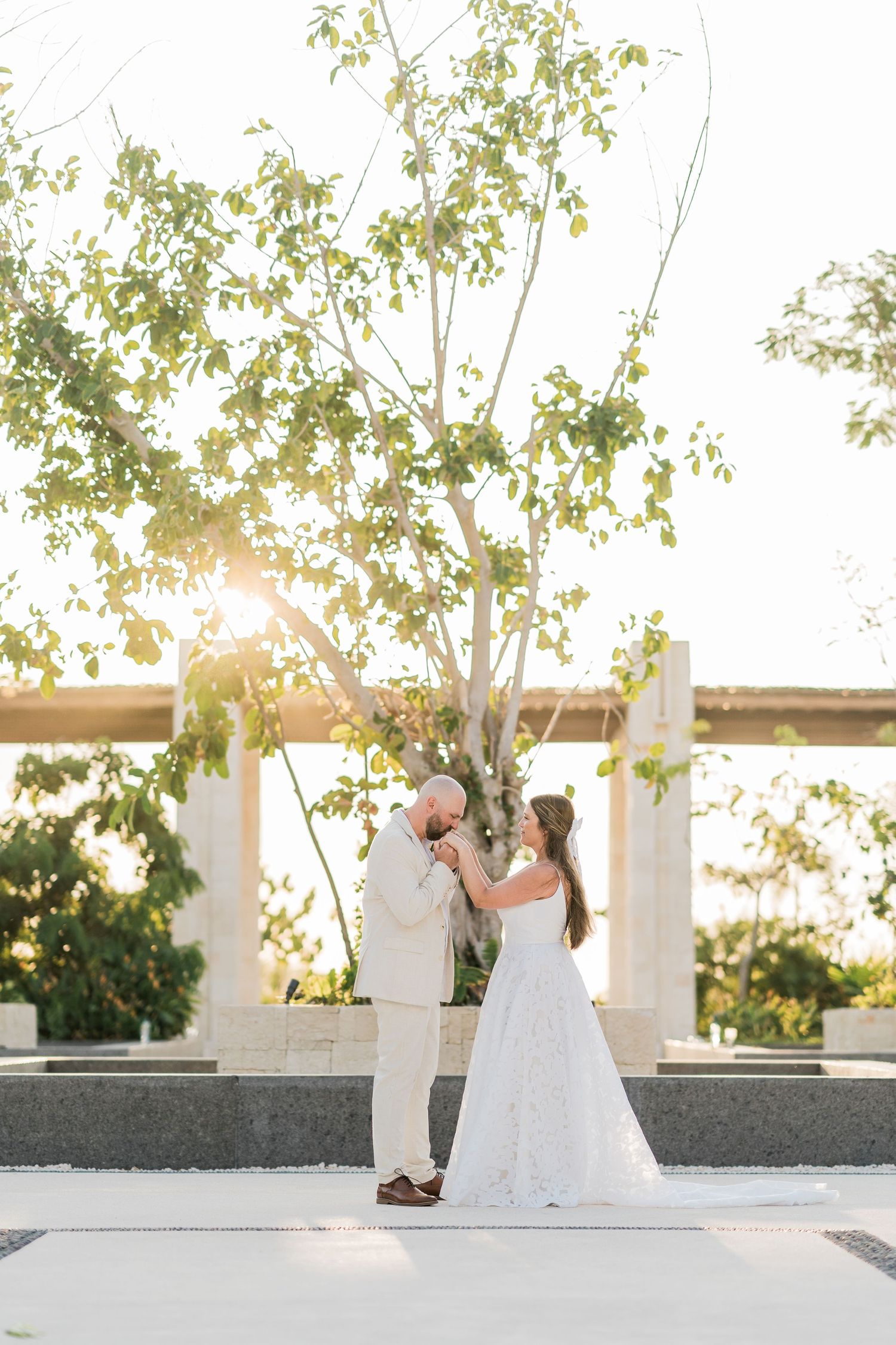 Bride and groom share a romantic moment under a sunlit tree at an outdoor wedding venue.