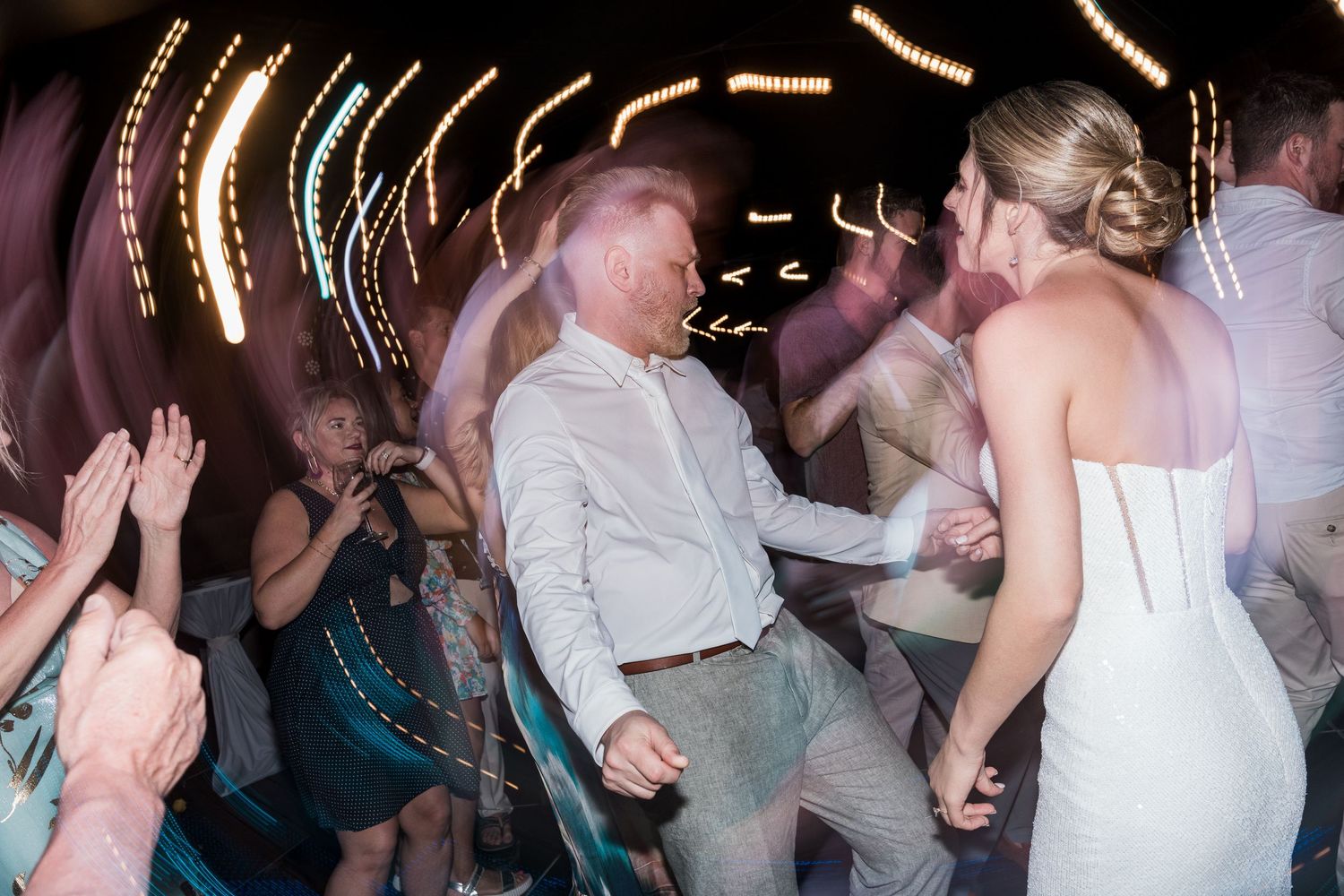 Bride and groom dancing joyfully with guests at a lively wedding reception under colorful lights.