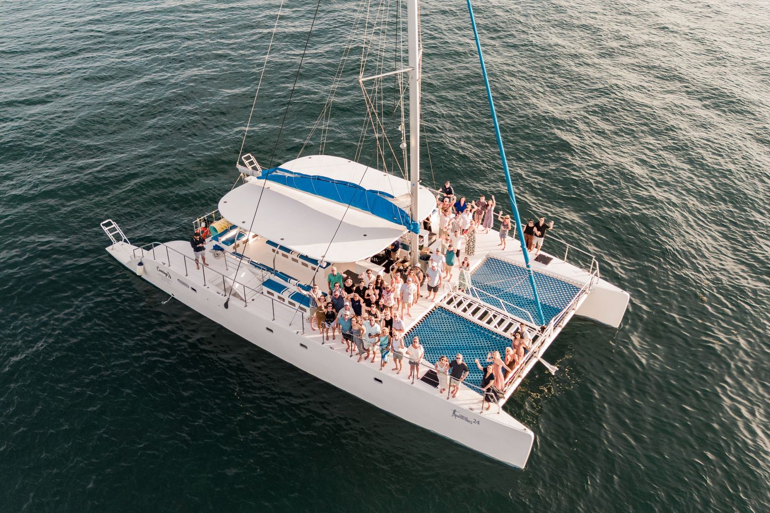 Group of people partying on a white catamaran sailboat on calm ocean waters during a sunset cruise.