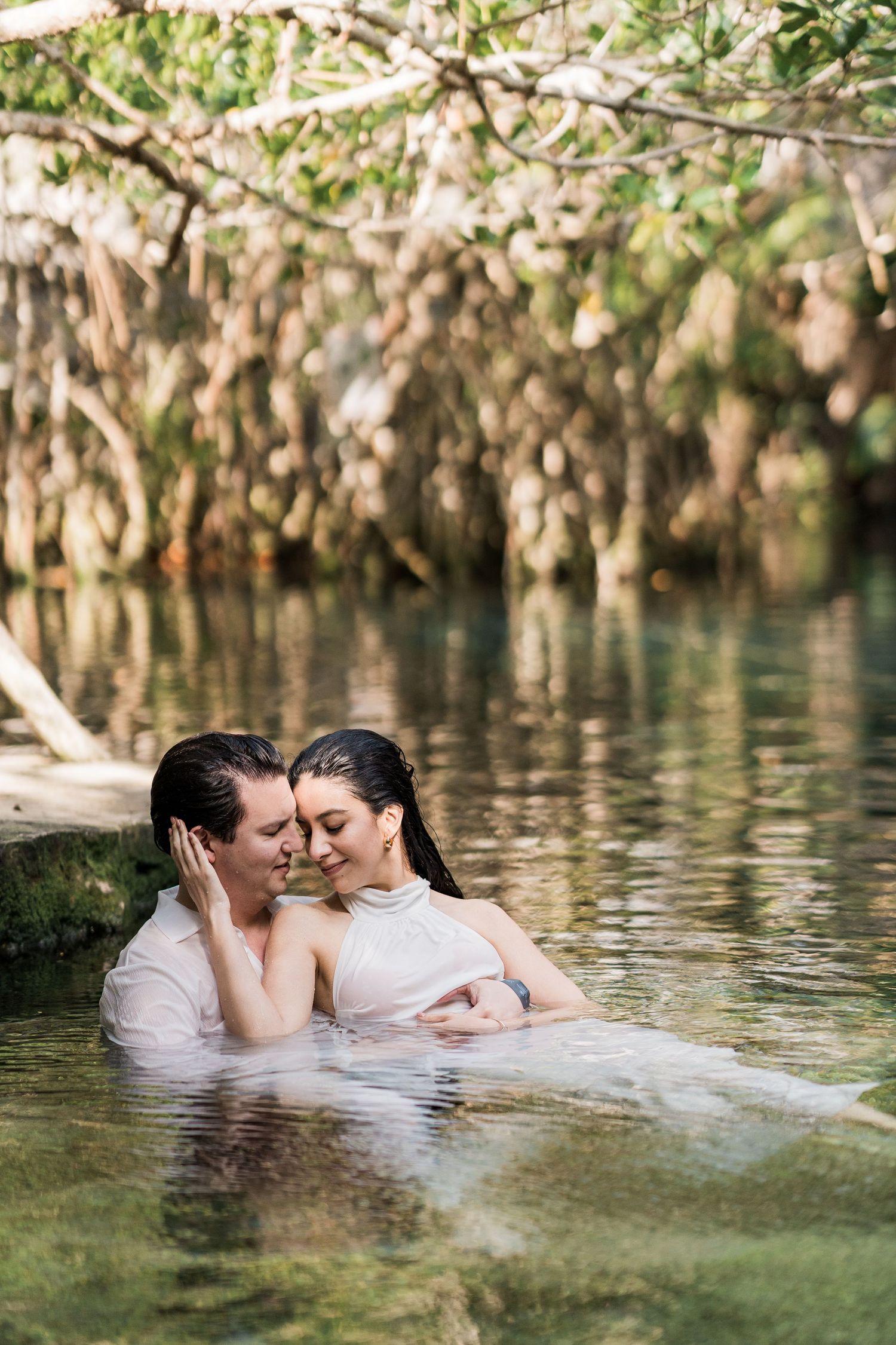 Couple in white attire embracing in a serene natural pool surrounded by lush tropical mangrove trees.