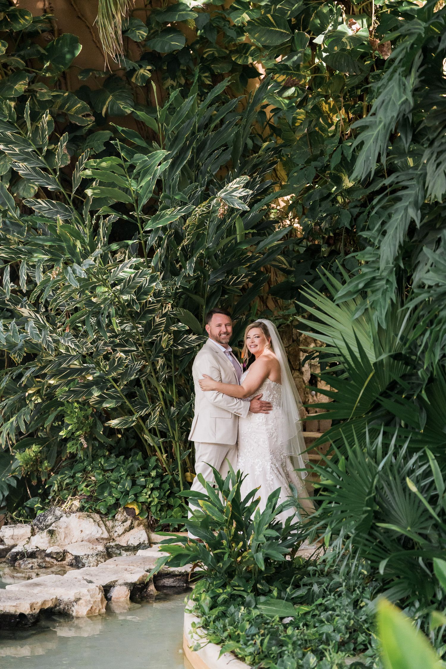 Bride and groom embracing among lush tropical greenery near a serene water feature on their wedding day.