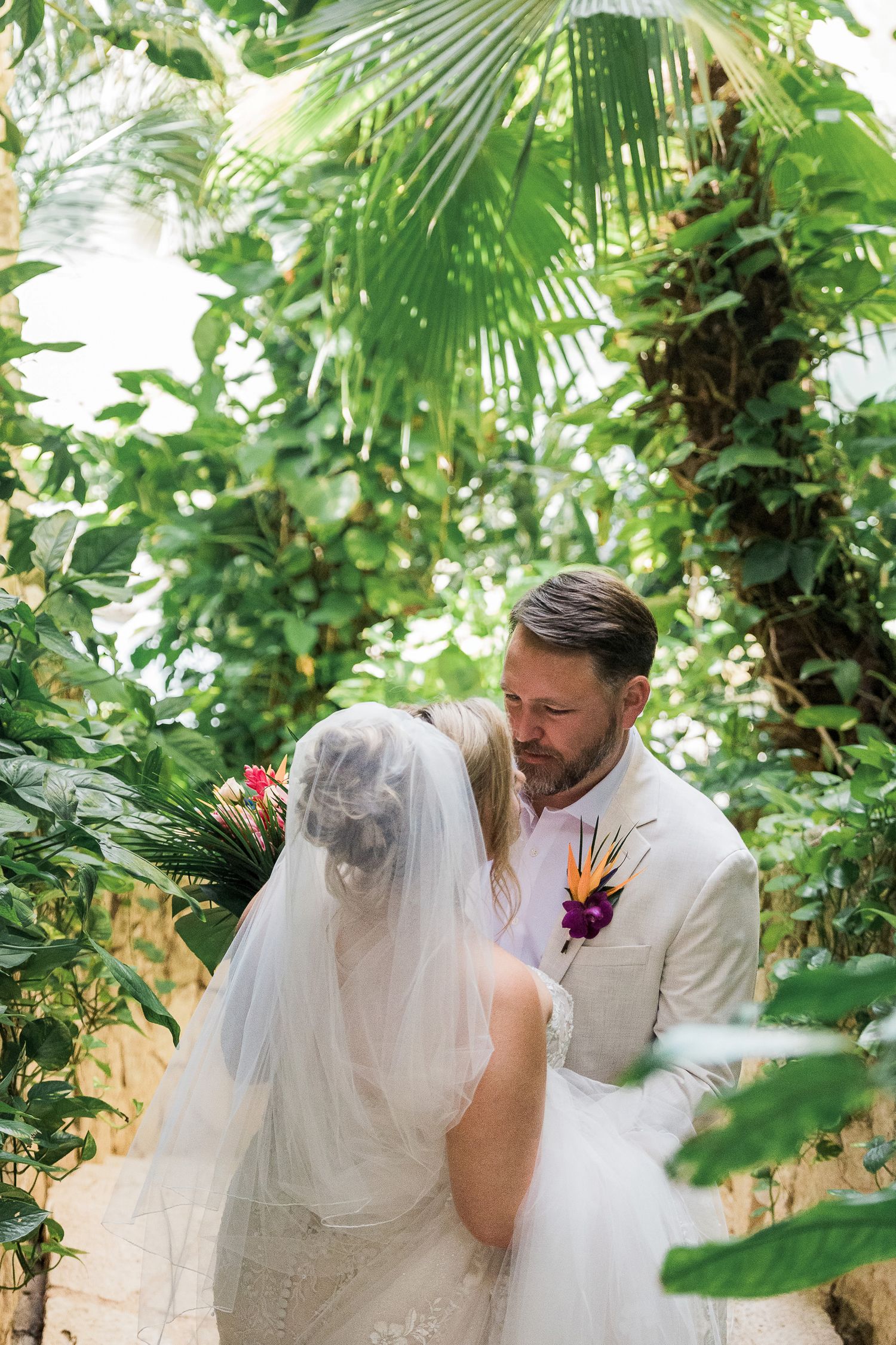 Bride and groom share intimate moment surrounded by lush tropical greenery, groom wearing beige suit with colorful boutonniere.