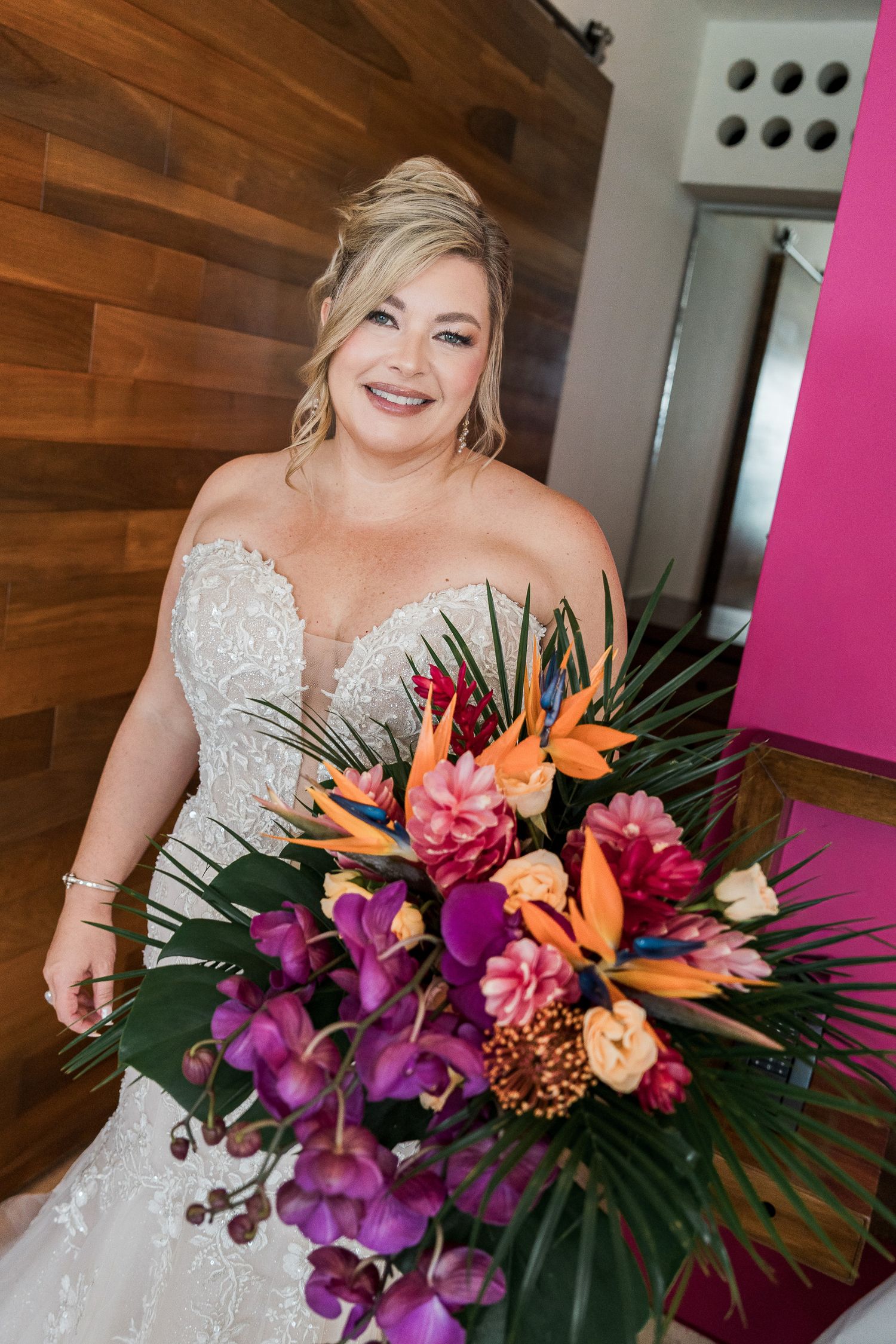 Smiling bride in lace gown holds vibrant tropical bouquet with birds of paradise, orchids, and palm leaves against wood wall.