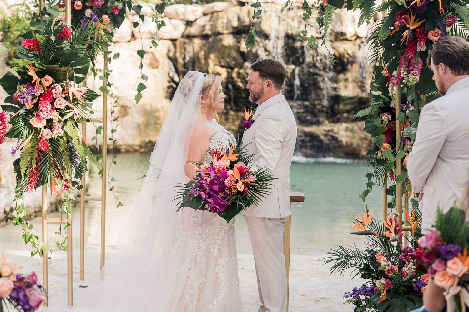 Bride and groom exchange vows under a floral arch with tropical blooms near a waterfall at an outdoor wedding ceremony.