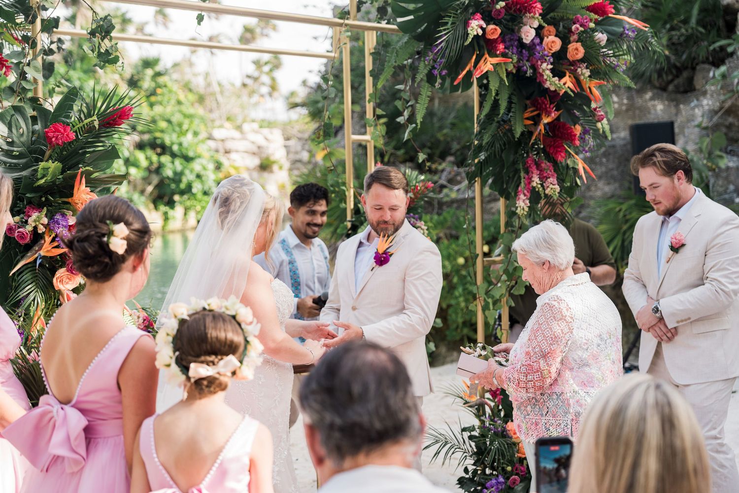 Outdoor wedding ceremony with couple exchanging vows under floral arch with colorful tropical flowers, guests in pink and beige attire.