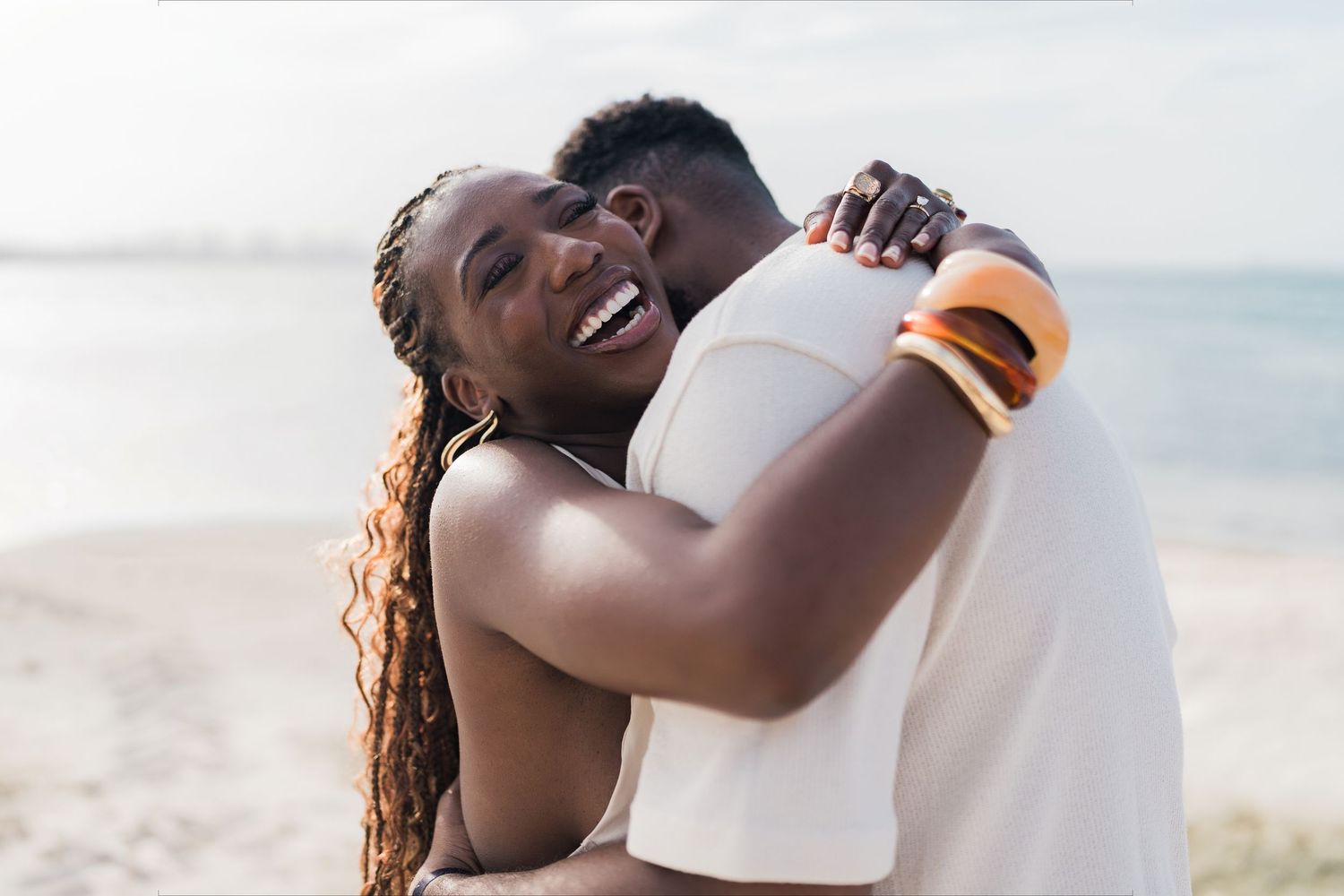 Happy couple embracing on a sunny beach, woman with long braids laughing joyfully while hugging her partner.